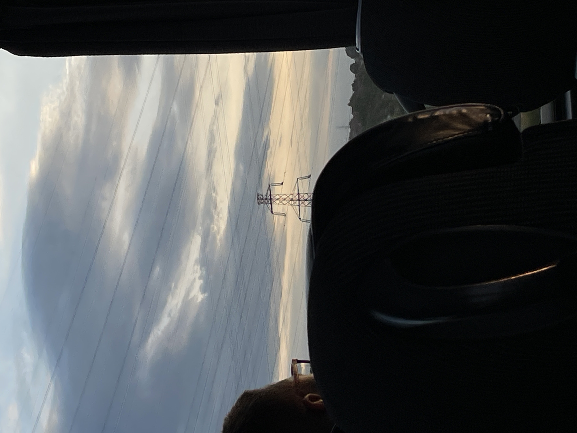 The image shows a view from inside a vehicle, likely a car or bus, looking out towards the sky. Overhead power lines and a utility tower are visible against a backdrop of cloudy skies, possibly indicating an overcast day. In the foreground are parts of the vehicle's interior, including a headrest and the top of a person's head, partially visible. The location is likely in the Surcin Urban Municipality, Serbia, due to the presence of the utility tower and overhead power lines, which are common infrastructure elements.