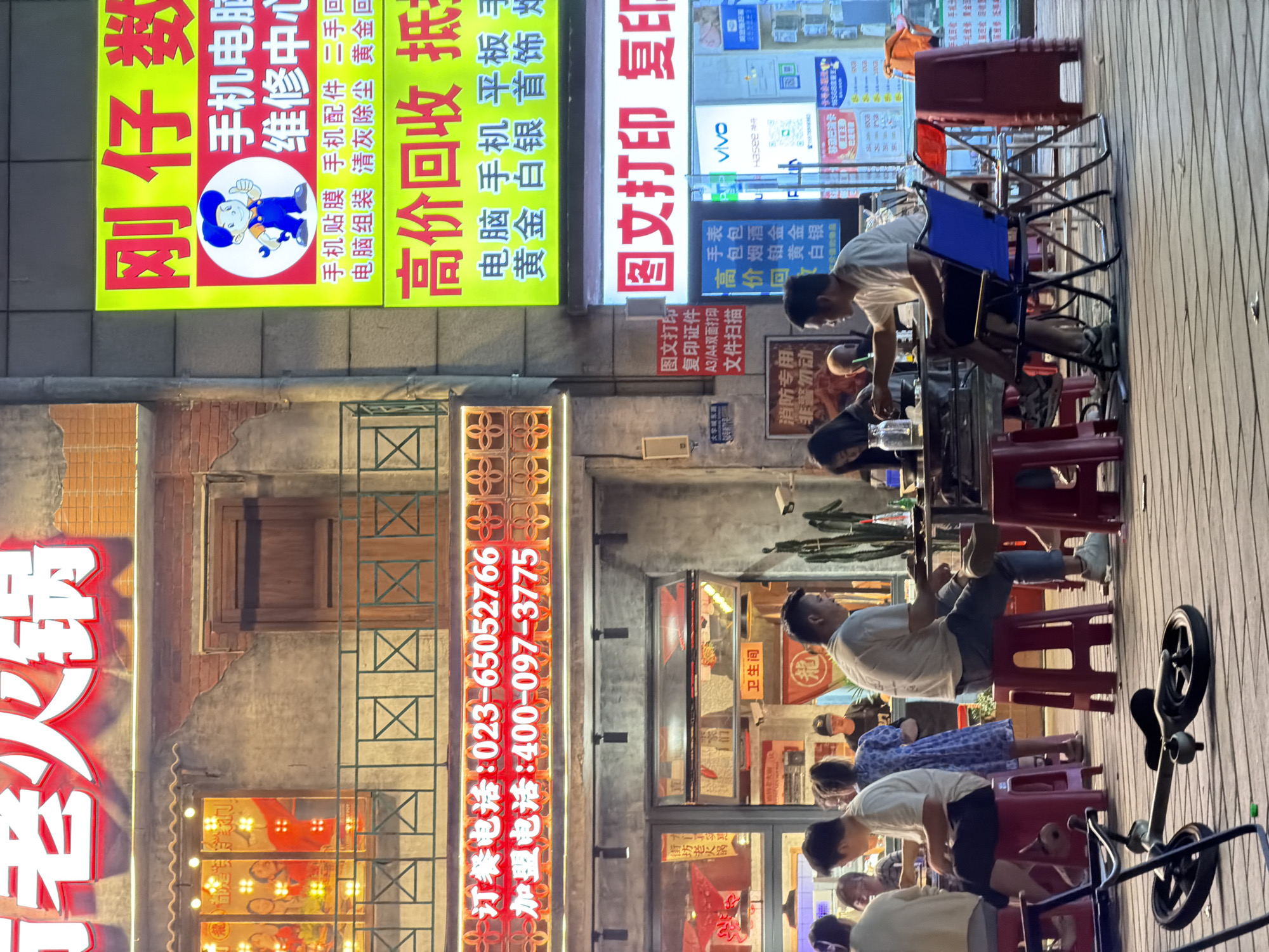 The image shows a street scene at night in the Chongqing High-tech Zone, China. Several people are seated on small red stools outside a restaurant. A few are seated around a table with clear plastic containers. The street is lined with buildings adorned with illuminated signs in Chinese characters. The signs indicate businesses, including a mobile phone repair shop, a printing shop, and a restaurant. The mobile phone repair shop sign has a cartoon character on it. The restaurant sign advertises hot pot and lists phone numbers. There is a metal grid in front of the restaurant, and windows display promotional images. Some chairs and a bicycle are placed on the sidewalk. The location is identifiable from the contact information on the sign.