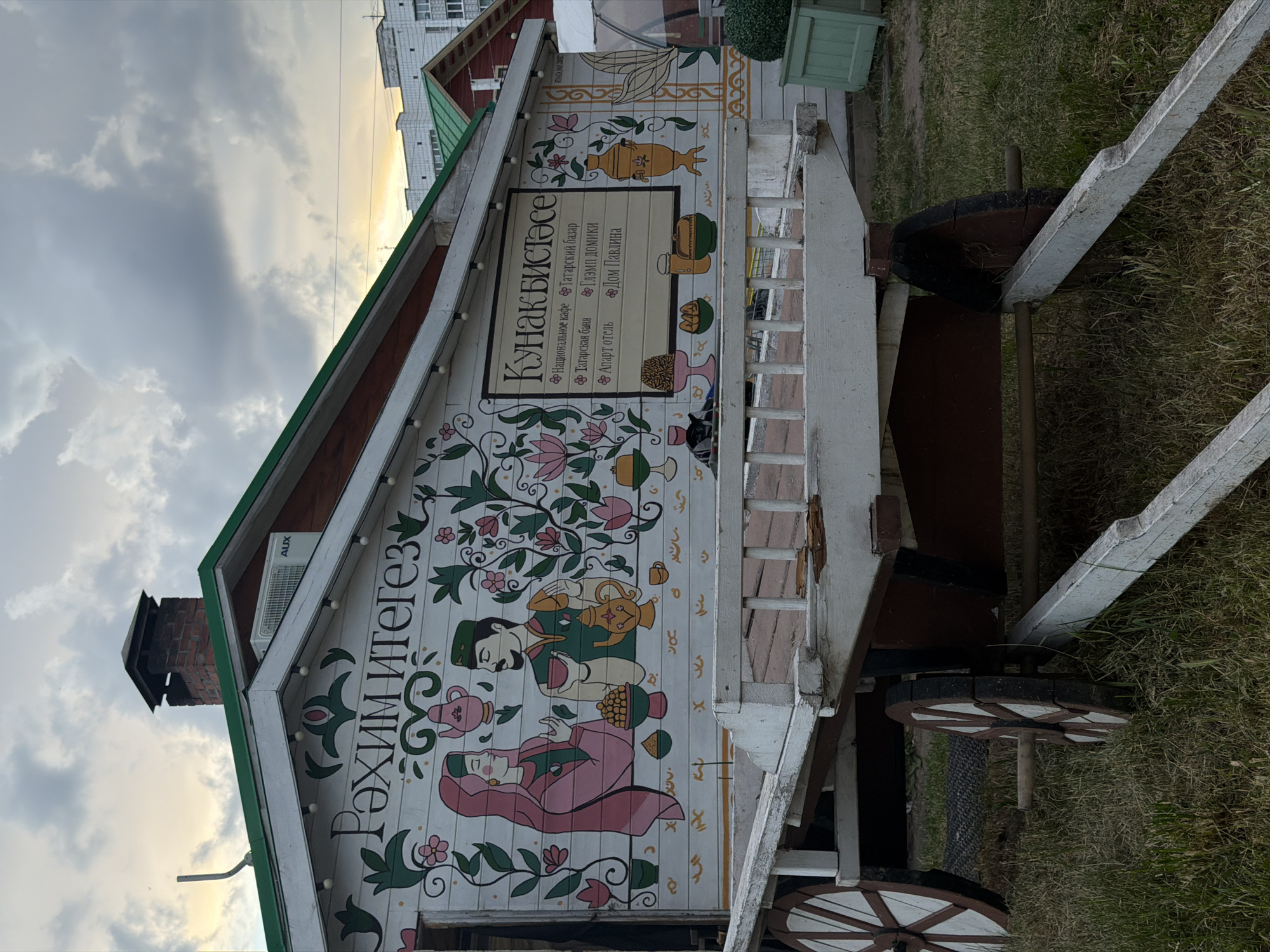 In an outdoor setting, a white wooden building with green trim features ornate folk art murals and signage. A prominent sign reads "Кунакбистəсе" in a decorative font, beneath which is a list of services: "Национальное кафе", "Татарская баня", "Апарт отель", "Татарский базар", "Глəмп домики", and "Дом Павлина". The murals depict stylized figures in traditional attire, teapots, flowers, and geometric patterns. In the foreground, a weathered white wooden cart with spoked wheels rests on a grassy area. The sky above is overcast with hints of sunset. An air conditioning unit is visible on the building's facade.
