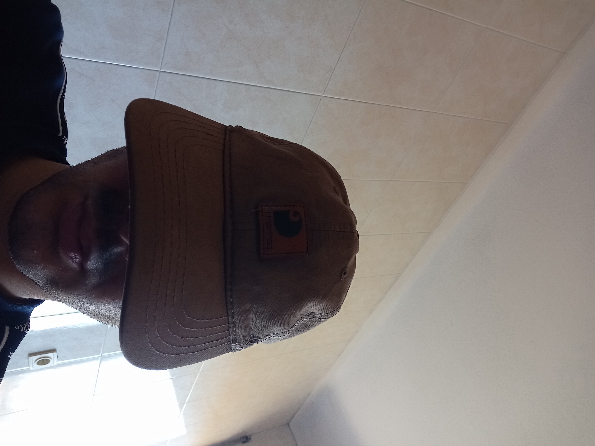 An individual is depicted from the mid-forehead downwards, wearing a brown baseball cap that obscures the upper half of the face. The cap features a brown rectangular patch with the "Carhartt" logo (visible in reverse, suggesting a mirror image or front-facing camera shot). The subject has stubble or short facial hair around the chin and mouth area, and is wearing a dark blue shirt with a patterned trim. The background consists of a wall covered in light beige square tiles with white grout lines. Above the tiled wall, a plain white ceiling is visible. To the right, a strong light source, possibly a window or doorway, creates a bright white area. Below this light source, a white electrical outlet or switch with a single circular button is visible on the tiled wall. The scene is located indoors, likely within a room such as a kitchen, bathroom, or utility area, in the city of Unknown, Unknown.