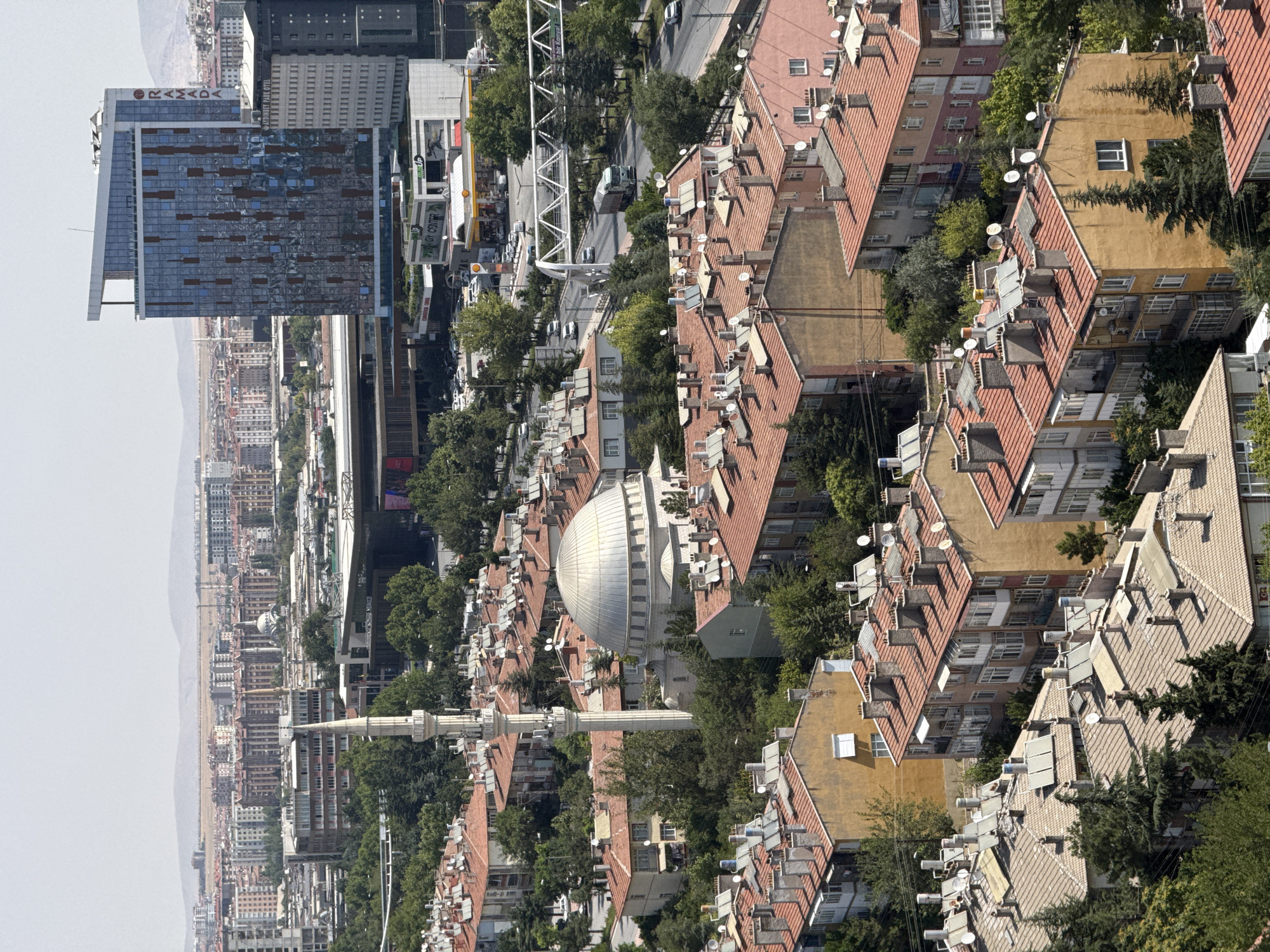 The image presents a panoramic view of a dense urban landscape in Konya, Turkey, characterized by a mix of residential buildings, modern architecture, and a prominent religious structure. In the foreground and midground, numerous multi-story apartment buildings with red-tiled roofs dominate the scene. Many of these roofs are equipped with solar water heaters and satellite dishes, indicating residential use and common amenities.
A distinctive feature of the midground is a mosque complex, featuring a large, silver dome and a tall, slender minaret. The mosque is nestled amongst the residential buildings and lush green trees. Further into the background, a modern, glass-fronted high-rise building stands out, bearing the "RAMADA" logo, suggesting a hotel or commercial establishment. Adjacent to it are other contemporary office buildings.
The horizon is lined with more distant buildings and a hazy, mountainous landscape, typical of an expansive city view. Roads with scattered vehicles, including a truck on a bridge, traverse the urban terrain. The overall atmosphere is one of a bustling city under a bright, possibly hazy, sky. There are no visible individuals in the frame, focusing the attention solely on the architectural and environmental elements of the city.
