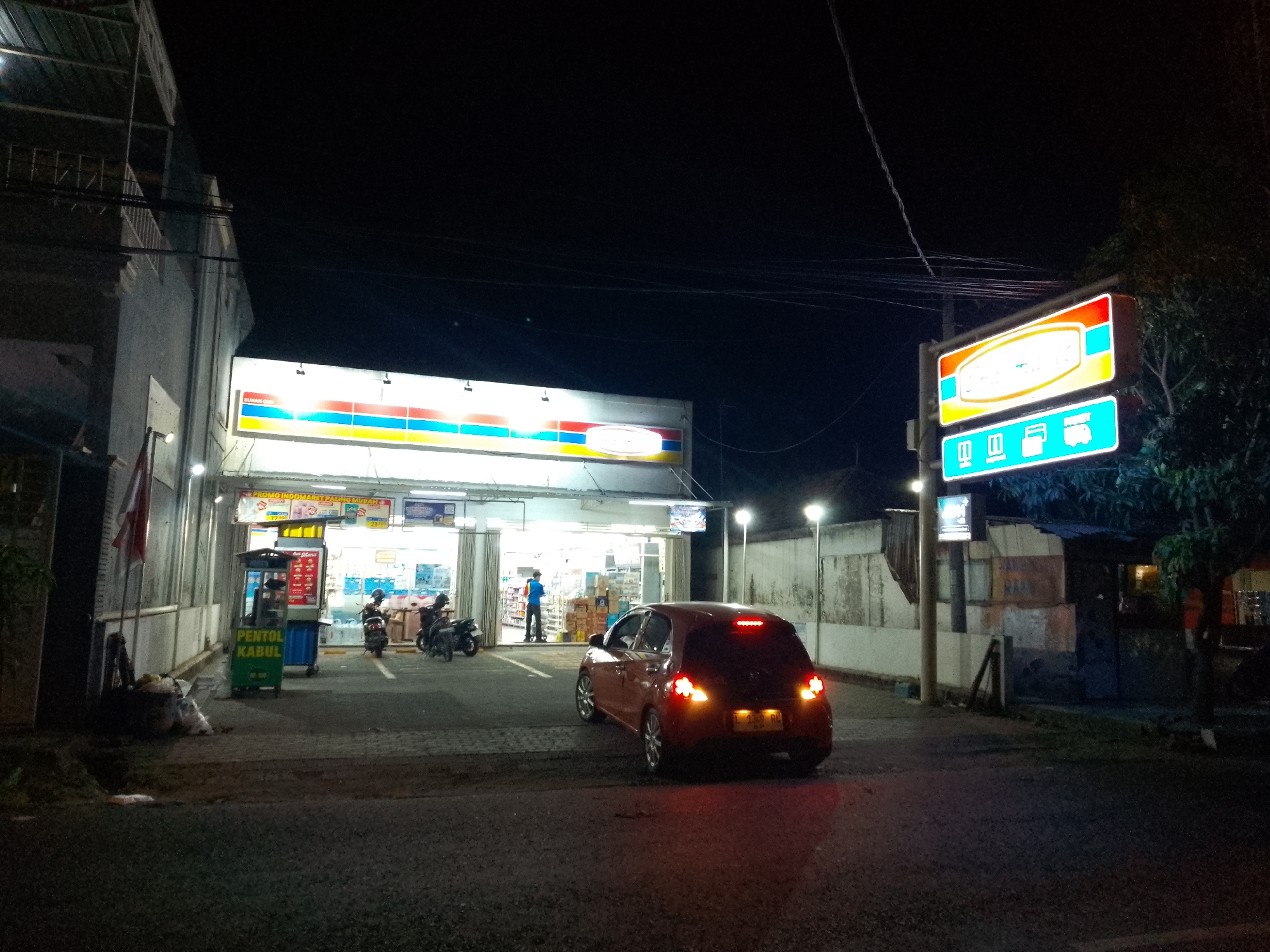 In the city of Lamongan, Indonesia, at night, a red hatchback car with its brake lights illuminated is parked in front of a brightly lit convenience store. The store has large glass doors and windows, with products visible inside. A sign above the store displays colorful horizontal stripes and a logo. To the right of the store, a prominent sign with an orange border and a light blue background features a rounded orange logo and several blue icons at the bottom, including what appears to be a credit card and a truck. A person in a blue shirt stands inside the store. Two motorcycles are parked near the entrance, and a small cart labeled "PENTOL KABUL" in green and blue is positioned to the left of the store entrance. The surrounding area is paved, with trees and a building with a metal roof visible in the background. Overhead wires are strung across the dark sky.