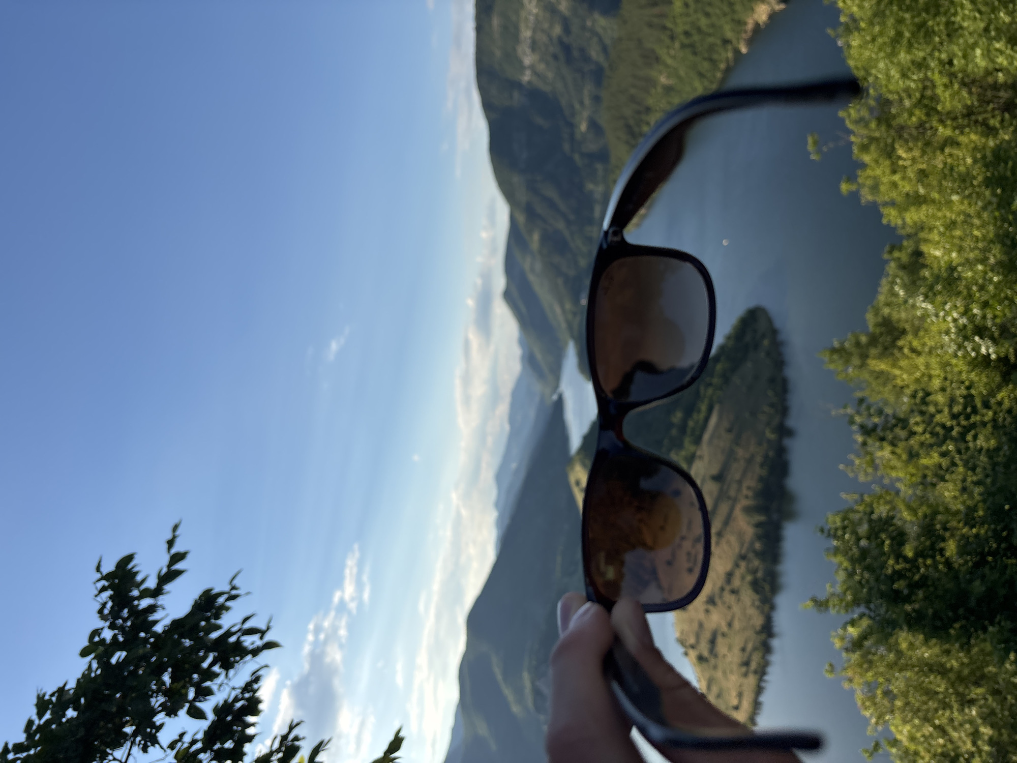 A close-up, over-the-shoulder perspective shows a hand holding a pair of dark-rimmed sunglasses in the foreground. The sunglasses are held at an angle, with their lenses partially obscuring a scenic view of a winding lake surrounded by lush, green, and rocky mountains. The sky above is a clear, bright blue with a few wispy clouds. The scene appears to be an outdoor landscape, possibly from a vantage point in Ribartsi, Bulgaria, given the mountainous terrain and the presence of a large body of water that could be the Studen Kladenets Reservoir, a popular natural attraction in the region. The sunlight suggests it is daytime, likely late afternoon given the gentle lighting. The focus is on the sunglasses and the partially visible landscape beyond them.