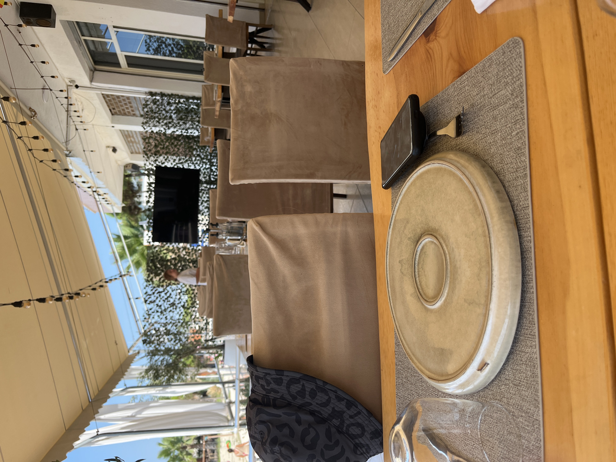 The image shows an outdoor seating area of a restaurant in Bashkia Durrës, Albania. The scene is set up for dining with tables and chairs. In the foreground, a wooden table is laid with placemats, a plate, a fork, a smartphone, and a glass. The table is set under an awning, with white curtains hanging to the side. In the background, more tables and chairs are visible, some covered with tan fabric. Palm trees and a beach area with people can be seen beyond the seating area. String lights are hung across the awning, and a television is mounted on a wall in the distance. The overall impression is a casual, beachfront dining experience.