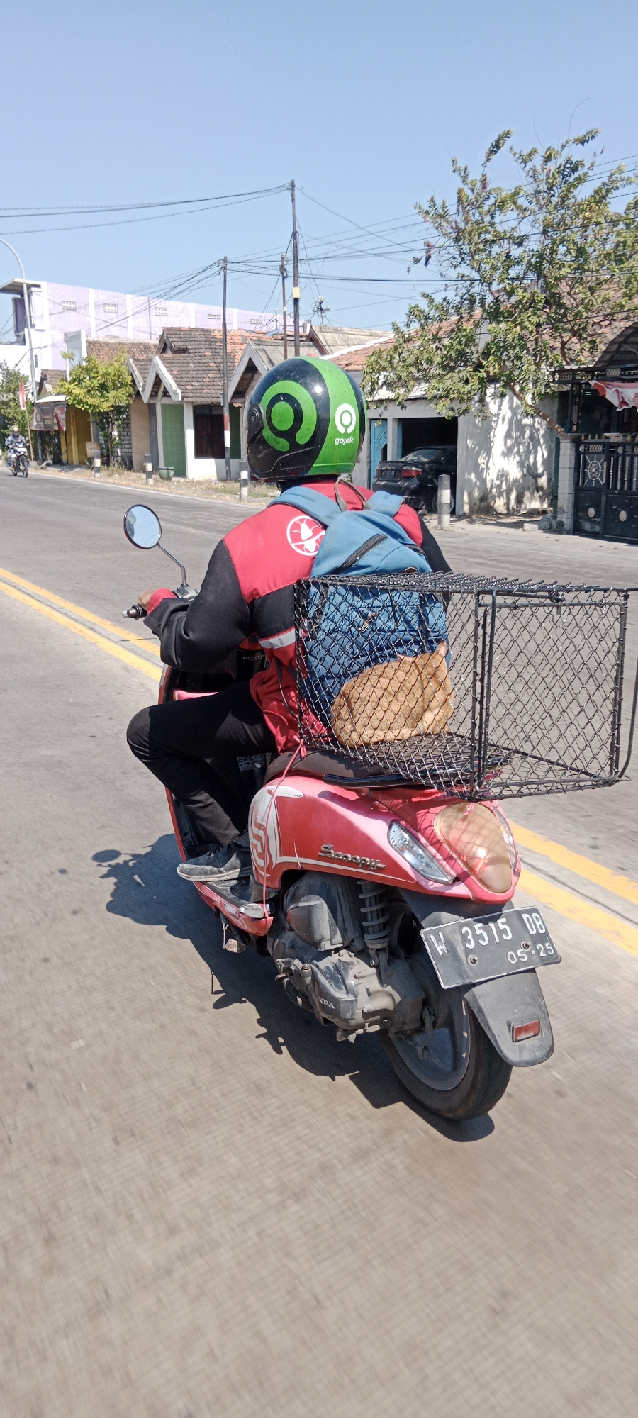 A person wearing a red and black jacket with a Gojek logo on the back and a green Gojek helmet is riding a red scooter. The scooter has a blue backpack attached to the rear rack, and a woven wire basket is mounted on top of the backpack. Inside the basket, a brown object, possibly an animal, is visible. The scooter is on a paved road with yellow lane markings. In the background, there are buildings with pitched roofs, trees, and utility poles under a clear blue sky. The license plate of the scooter reads "W 3515 DB 05-25". The scene appears to be in Samirplapan, Indonesia, based on the contextual information provided.