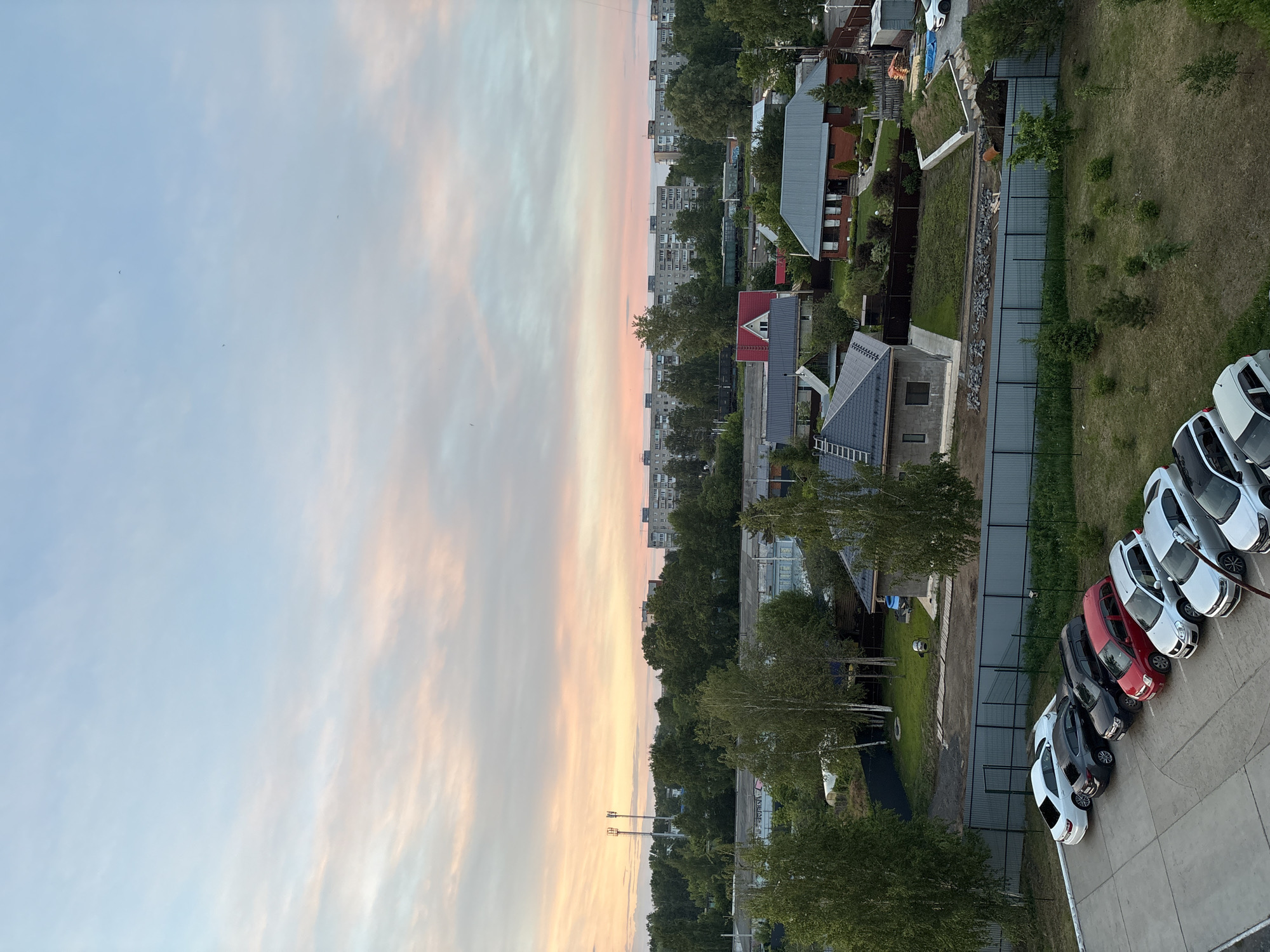 The image shows an aerial view of a cityscape at dawn or dusk. The sky is filled with light blue and pink clouds.
To the right, there are houses and buildings, with a mix of residential areas. There is also a parking lot with several cars parked.
The location is Chaikovsky, Russia.