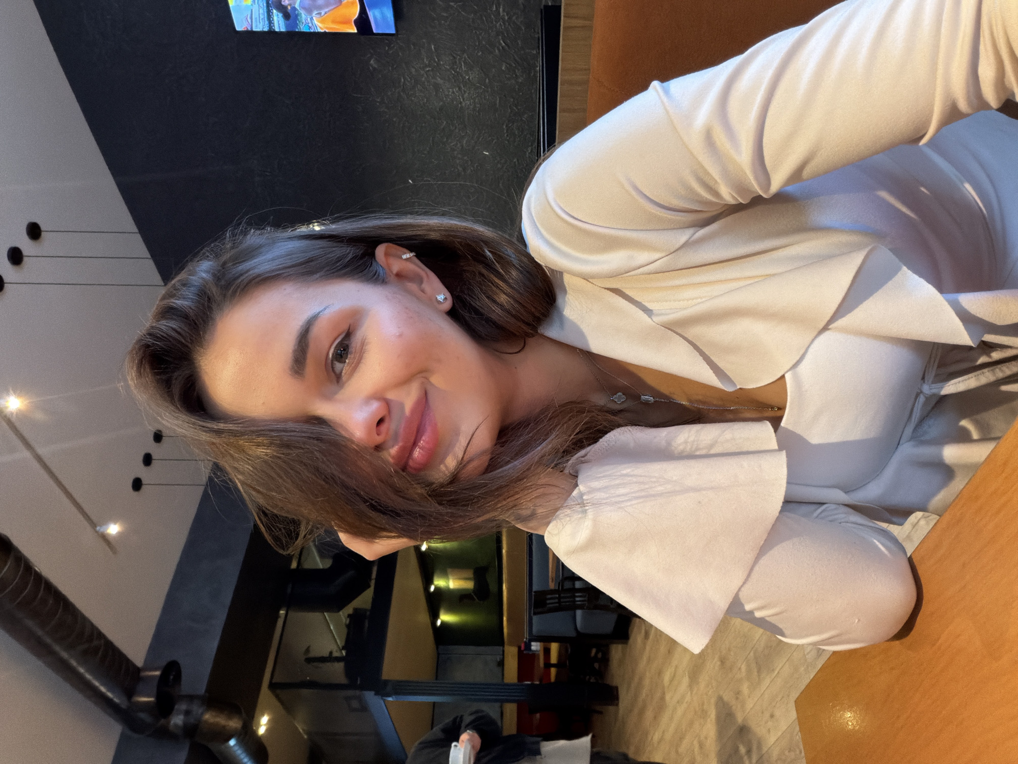 The image shows a woman sitting at a table, likely indoors. She has brown hair and is wearing a white blouse or dress. Her hair is partially covering her face. She appears to be smiling. The background features a dark wall, a table or counter, and some lighting fixtures. Part of a television screen is also visible in the background. The location is possibly Tbilisi, Georgia.