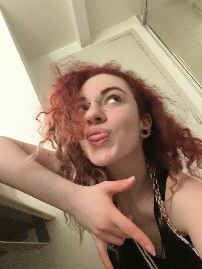 The image is a low-angle, close-up portrait of a young adult female with voluminous, curly red hair, captured indoors. She gazes upwards and to the right with a playful expression, her tongue extended slightly from pursed lips. Visible piercings include a silver barbell-style bridge piercing between her eyebrows and black tunnel plugs in her earlobes. She wears a black sleeveless top, and light-colored braided hair extensions are visible over her left shoulder. Her left hand is in the foreground, forming a gesture with the thumb and pinky finger extended while the middle and ring fingers are curled. Her fingernails are manicured with a clear or light polish. The background features plain, light-colored walls and ceiling. A vertical door frame or panel is partially visible in the upper right. A light-toned horizontal surface, possibly a counter or shelf, with a dark recessed area beneath it, is present in the lower left. No visual information within the image permits identification of a specific location in Paris, France.
