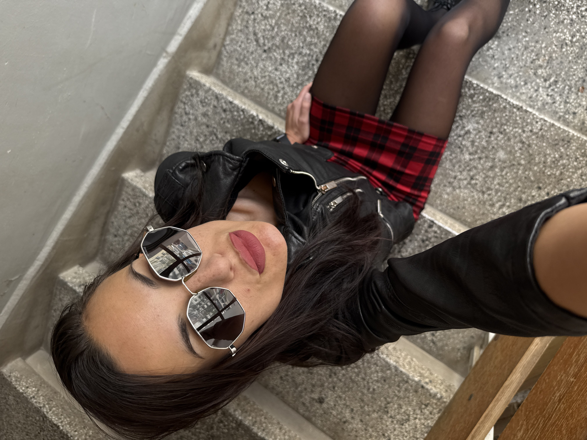 A young woman wearing a black leather jacket, red plaid mini skirt, black pantyhose, and silver-framed sunglasses lies on gray concrete steps. The location is inside a building with stairs. The geographic coordinates are not possible to determine from the image. Pleven, Bulgaria cannot be confirmed from the provided information.