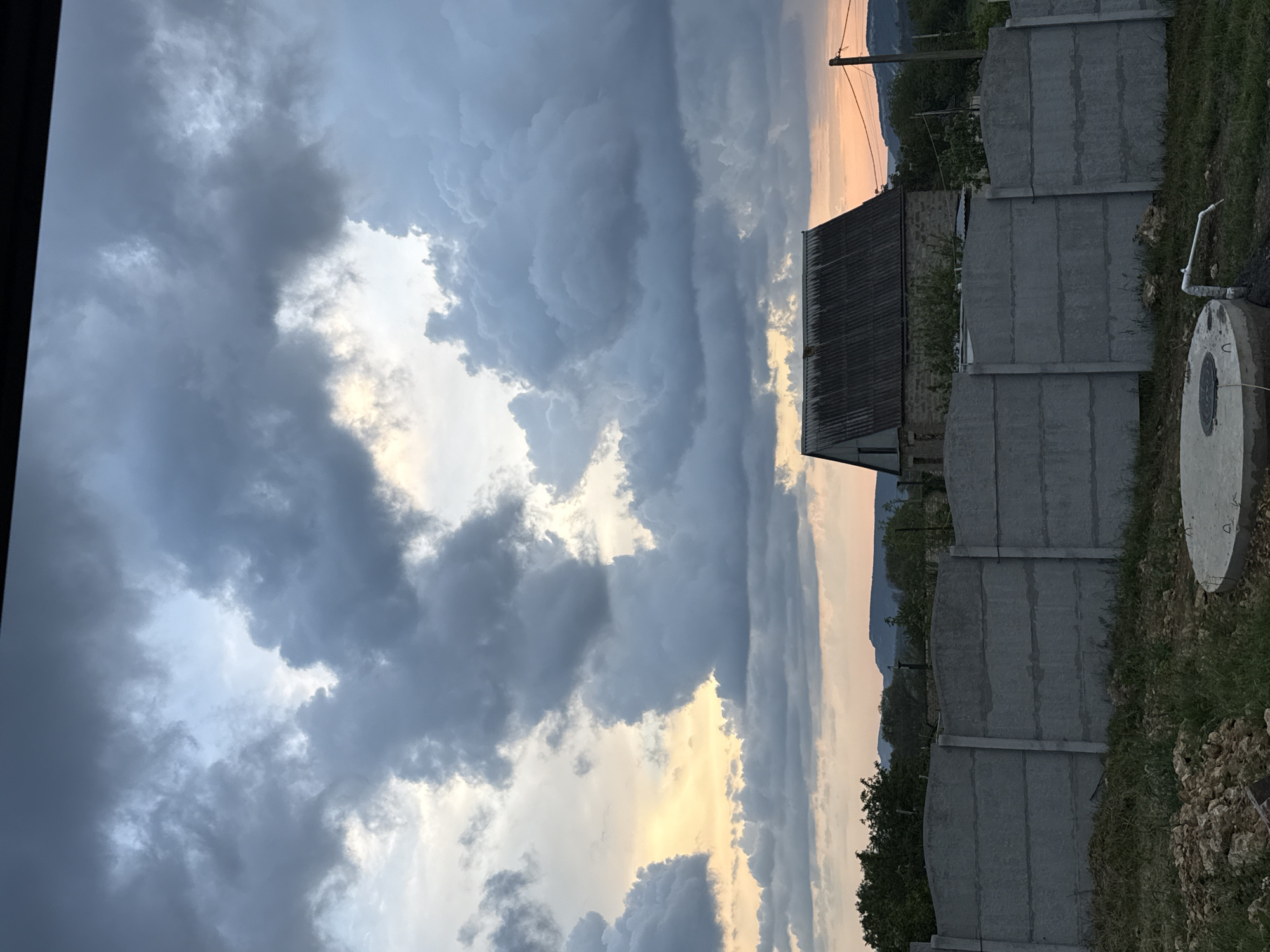 The image shows a view in Перовское сельское поселение, Ukraine, capturing a dramatic sky filled with heavy, dark clouds, some tinged with gold from the setting sun. Below the clouds, there's a glimpse of land featuring a low building with a metal roof, a concrete fence comprised of modular segments, and some vegetation. A utility pole with lines is also visible. A concrete well ring sits near the fence. The photograph is taken from a low angle and rotated counter-clockwise.