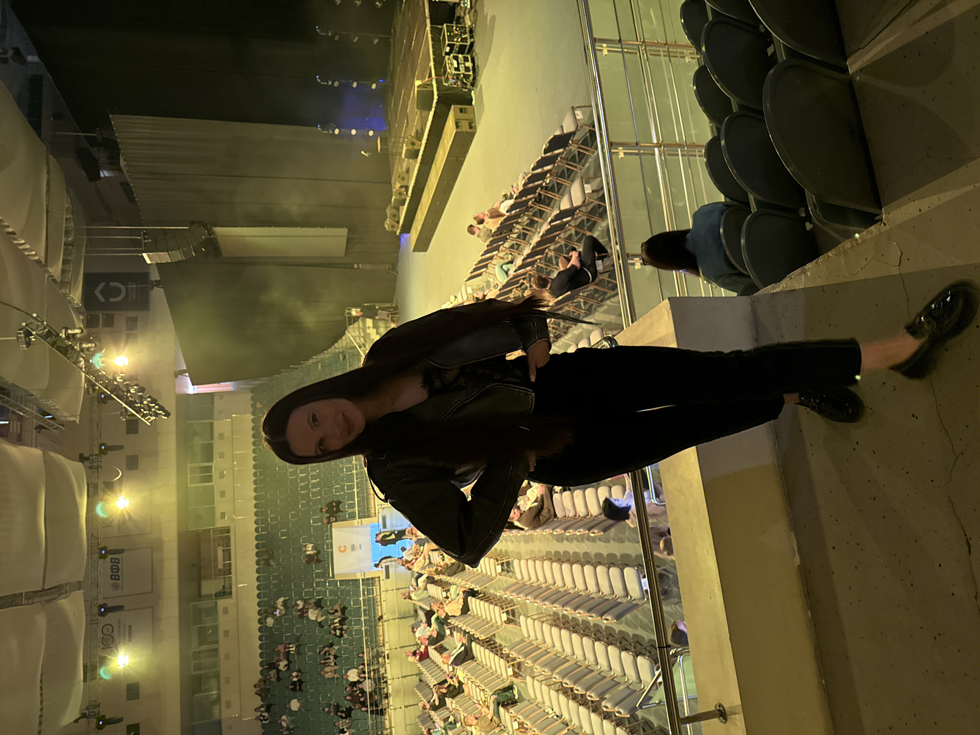An indoor, vertically-oriented photograph captures a large arena or event hall, likely during an evening event. The primary subject is a young woman, positioned on the right side of the frame, smiling directly at the camera. She has long, dark hair and is dressed in a dark leather-style jacket, dark top, dark trousers, and dark flat shoes. She appears to be standing or leaning against a concrete barrier or wall in an elevated seating section.
In the background, a large stage or platform is visible in the distance, equipped with various lighting fixtures and speakers. Rows of tiered seating, characteristic of an auditorium or stadium, extend throughout the venue. Many seats are occupied by an audience, primarily facing towards the stage, though some sections appear less populated. The overall lighting is artificial, with stage lights casting blue and white hues, and general venue lighting providing a yellowish-green ambient glow in certain areas.
Notable details include a concrete railing or barrier in the foreground, separating the woman from the lower seating sections. The atmosphere suggests a live performance, concert, or conference.
Several pieces of text and logos are visible in the background:
- A large rectangular panel displays "ВФВ" (likely Cyrillic characters) and possibly "BDB".
- Another black panel features a white stylized logo resembling `<O>` or `<C>` with an arrow.
- A banner in the mid-ground shows an orange circular logo with an incomplete "C" shape.