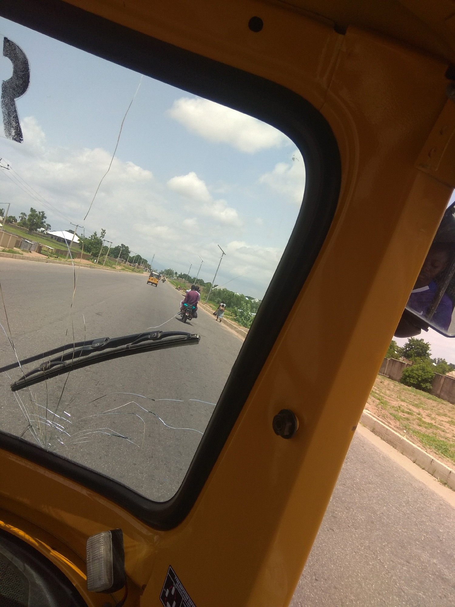 The image, taken from inside a yellow vehicle in Jalingo, Nigeria, shows a cracked windshield with the road and its surroundings visible through it. The road is paved and appears to be a main thoroughfare, with a yellow motorized tricycle, commonly known as a "Keke Napep," traveling in the distance. A motorcycle with two riders is also visible on the road. The environment is somewhat rural, with trees and buildings lining the roadside, and a clear sky with scattered clouds above. A black object, possibly a steering wheel, is partially visible in the lower left corner of the frame, and a wiper blade is present on the windshield. The overall impression is of a journey through a town in Nigeria.
