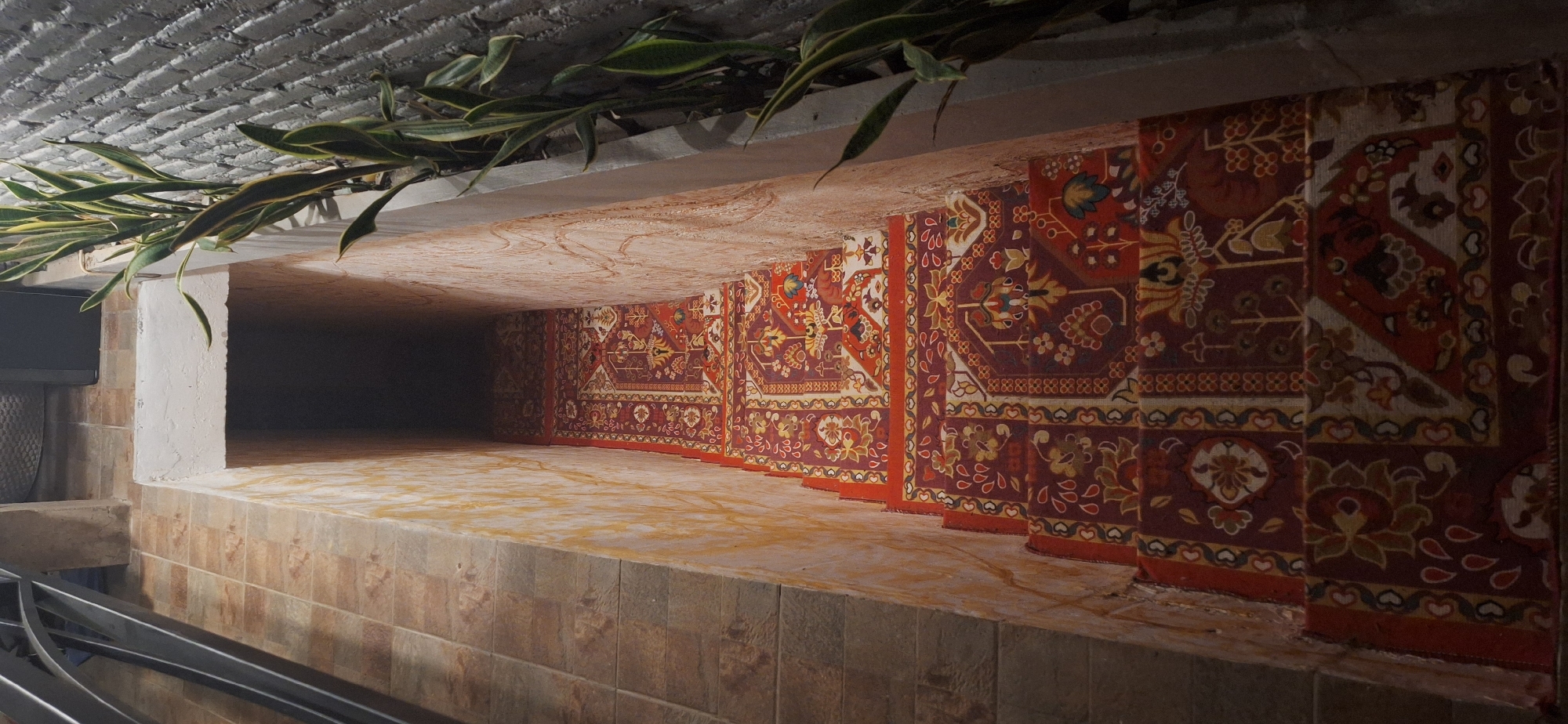 The image shows a narrow, descending staircase in Kuta Selatan, Indonesia. The stairs are covered with a richly patterned Persian-style carpet in shades of red, maroon, yellow, and blue. The walls of the staircase are textured, with the left wall appearing to be made of large, rectangular stone tiles and the right wall exhibiting a rough, plaster-like finish with hints of yellow and orange marbling. A tall, leafy plant, possibly a snake plant, grows along the right side of the staircase, adjacent to a textured grey brick wall. The top of the staircase is partially visible, with a concrete lip and a metal railing on the left. The overall scene suggests an outdoor or semi-outdoor area, perhaps a pathway or entrance to a building. There are no people visible in the image.
