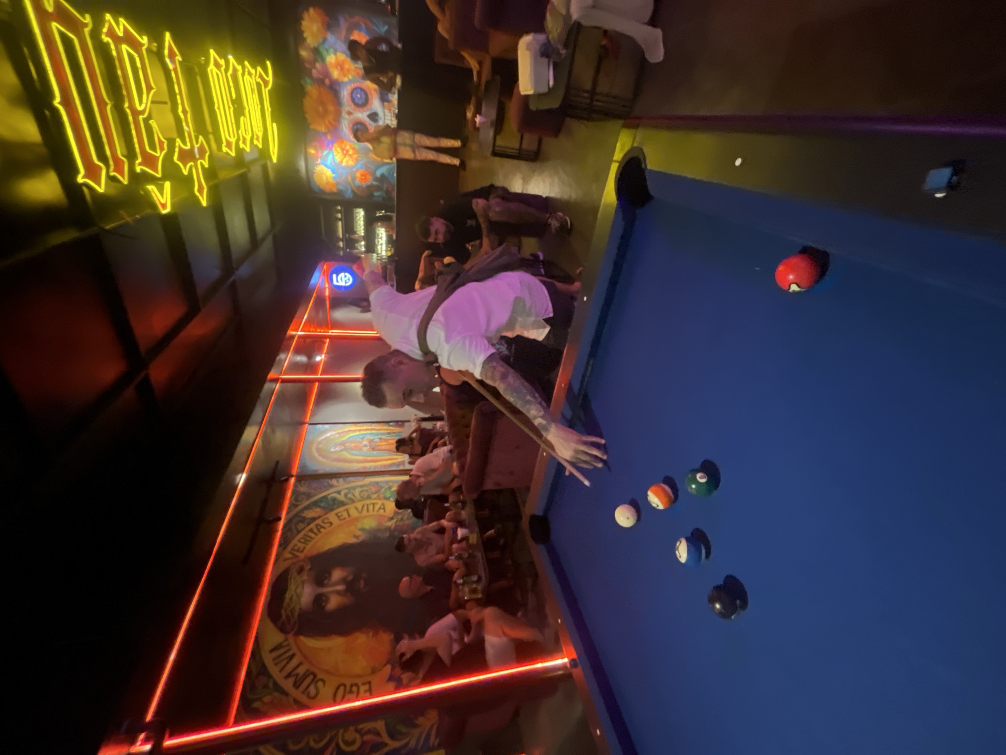 The image depicts a dimly lit interior, possibly a bar or lounge, with vibrant neon lighting and large murals adorning the walls. In the foreground, a person is actively playing pool on a blue felt table. Several pool balls are visible on the table, including a black ball, a blue and white striped ball, a green ball with the number 6, and a red ball with the number 7 near a corner pocket. The player, dressed in a white shirt and dark shorts, is in the process of taking a shot, holding a pool cue.
In the background, other patrons are seated at tables and on couches, engaged in conversation and enjoying drinks. The walls feature colorful murals, one depicting religious imagery with the text "EGO SUM VIA VERITAS ET VITA," and another with a skeleton motif often associated with Day of the Dead celebrations. The overall atmosphere suggests a lively social gathering. The location is not identifiable as Đà Nẵng, Vietnam, based solely on the visual information provided.