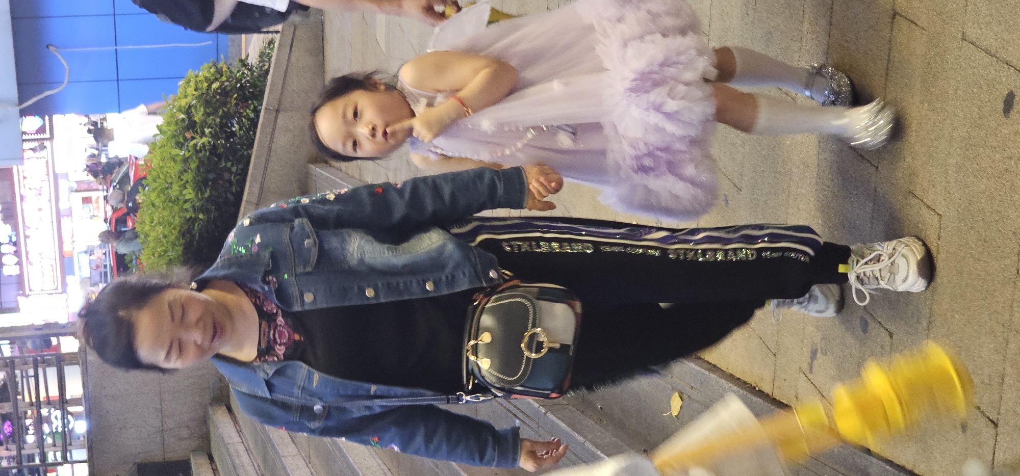 A woman and a young girl are standing outside on a sidewalk in what appears to be an urban area. The woman is on the left, wearing a denim jacket with colorful embellishments, a black top, black pants with a reflective stripe, white sneakers, and a crossbody bag. The young girl is to the right of the woman, wearing a light purple dress, white socks, and sparkly silver shoes. The girl is holding a finger to her lips. In the background are plants, storefronts, and signs. The location is in the city of Yuzhong District, China.