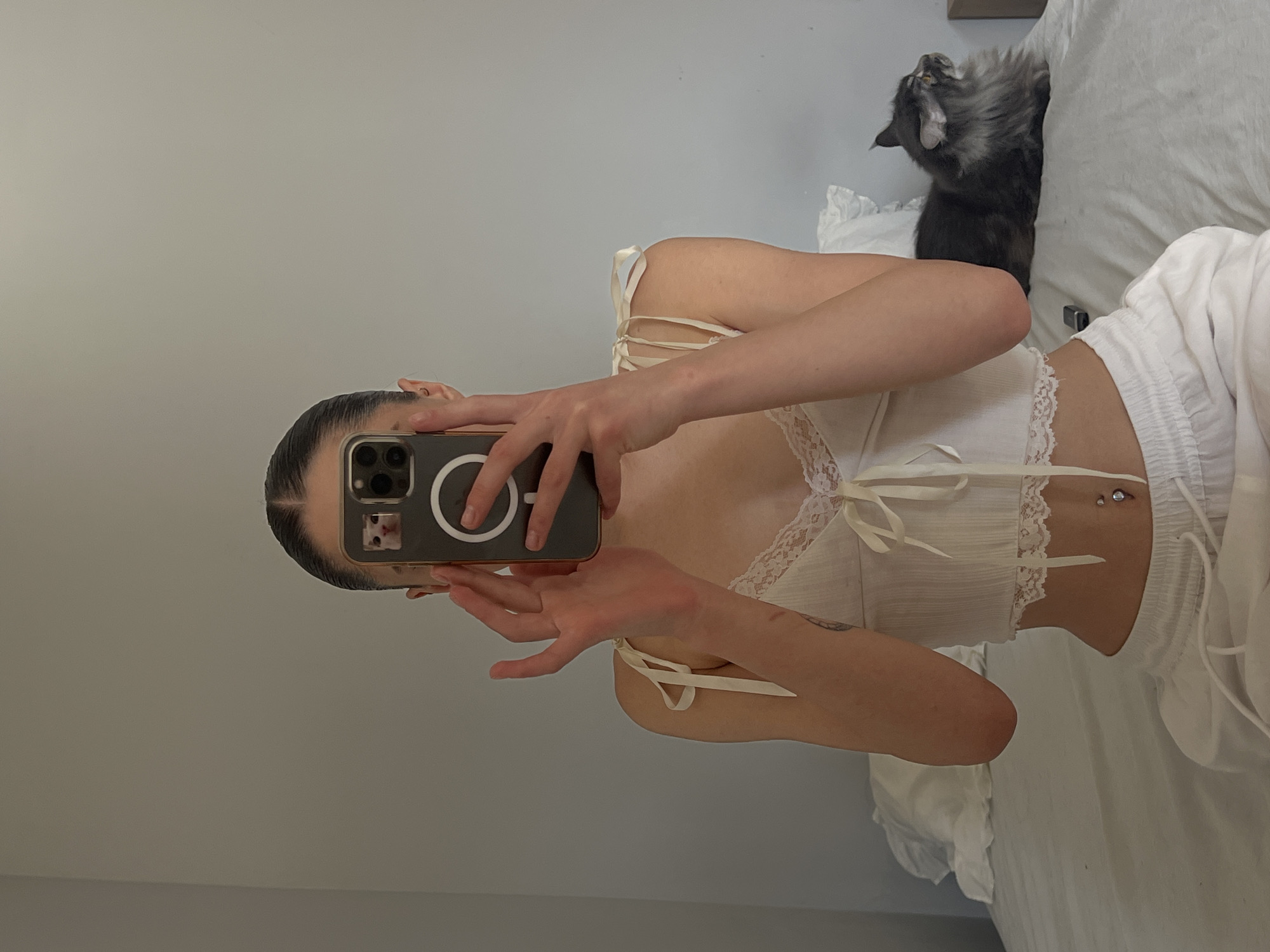 In a dimly lit bedroom, a young woman, seen from the chest up, is taking a mirror selfie. She has her hair pulled back tightly, revealing a simple ear piercing. She is wearing a white camisole with lace trim and ties at the shoulder straps. A silver belly button ring is visible. To her right, a long-haired gray cat is lying on a white bed. The room appears to be sparsely decorated, with light blue walls and white bedding. The overall atmosphere is intimate and casual. There are no indications that this scene is located in Ternopil, Ukraine.