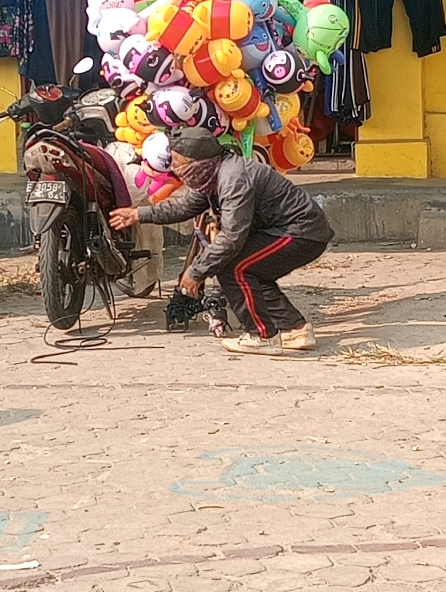A person wearing a mask and a dark grey jacket with red stripes down the sleeves is squatting next to a motorcycle. The person is reaching down towards a black cable on the ground. A bundle of colorful inflatable toys, including cartoon characters and balloons, is attached to the motorcycle. The motorcycle has a license plate that reads "E 3058 B". The scene appears to be on a paved street with a yellow wall in the background. The location is specified as Bandar Lampung, Indonesia.