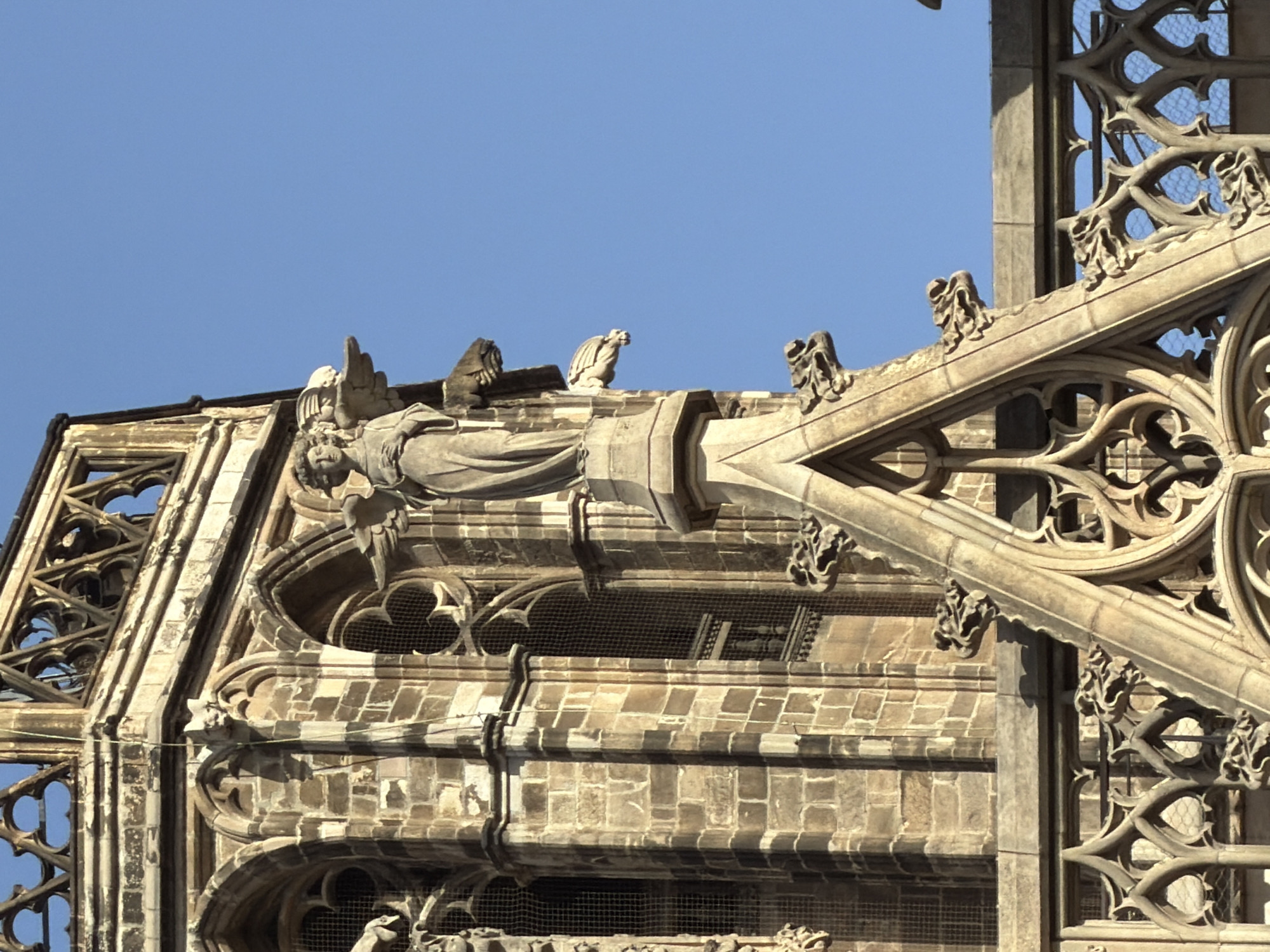 The image shows a close-up view of the ornate Gothic architecture of a cathedral. A stone statue of an angel with feathered wings is prominently featured on the upper portion of a spire. The angel is depicted in a flowing robe, with its hands clasped in front. The statue is weathered, indicating its age. Below the angel, the spire continues with intricate stone carvings, including gargoyles, which are grotesque carved figures. The background is a clear, bright blue sky. The architectural style suggests it is likely part of a historical cathedral, possibly in Europe. The stonework is detailed and features pointed arches, decorative tracery, and weathered surfaces, all characteristic of Gothic cathedrals. The detailed stonework and the presence of gargoyles are common features of such structures. Without further context or identifiable landmarks, pinpointing the exact location within Barcelona, Spain, is not possible from the image alone.