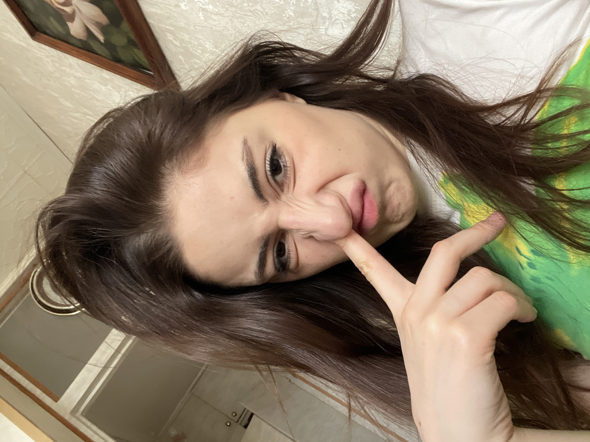 A woman is indoors. She has long, dark hair and is wearing a white and green shirt. She is holding her finger near her nose and making a funny face. There is a painting on the wall and part of a doorframe visible. The location is not discernible as Moscow, Russia, from the image.