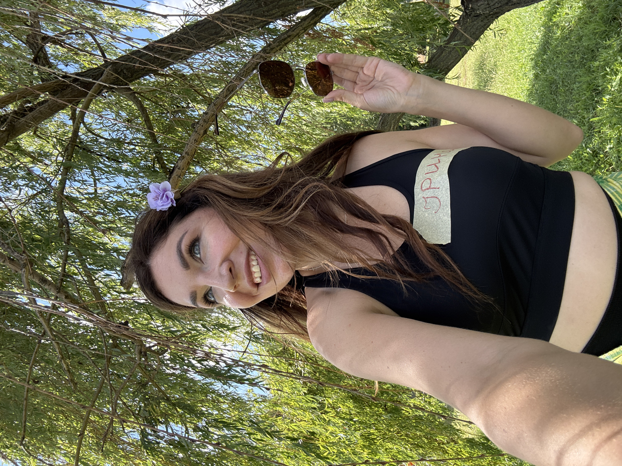In the city of Mar'yanivka, Ukraine, a woman with long, brown hair adorned with a light purple flower stands outdoors. She is wearing a black sports bra with a beige band that has red Cyrillic lettering written on it. She is holding a pair of sunglasses in her right hand. The woman is smiling and looking upwards and to her left. She is standing under a large tree with weeping branches, and the background consists of green grass and a blue sky with some clouds. The lighting suggests it is daytime, likely sunny.