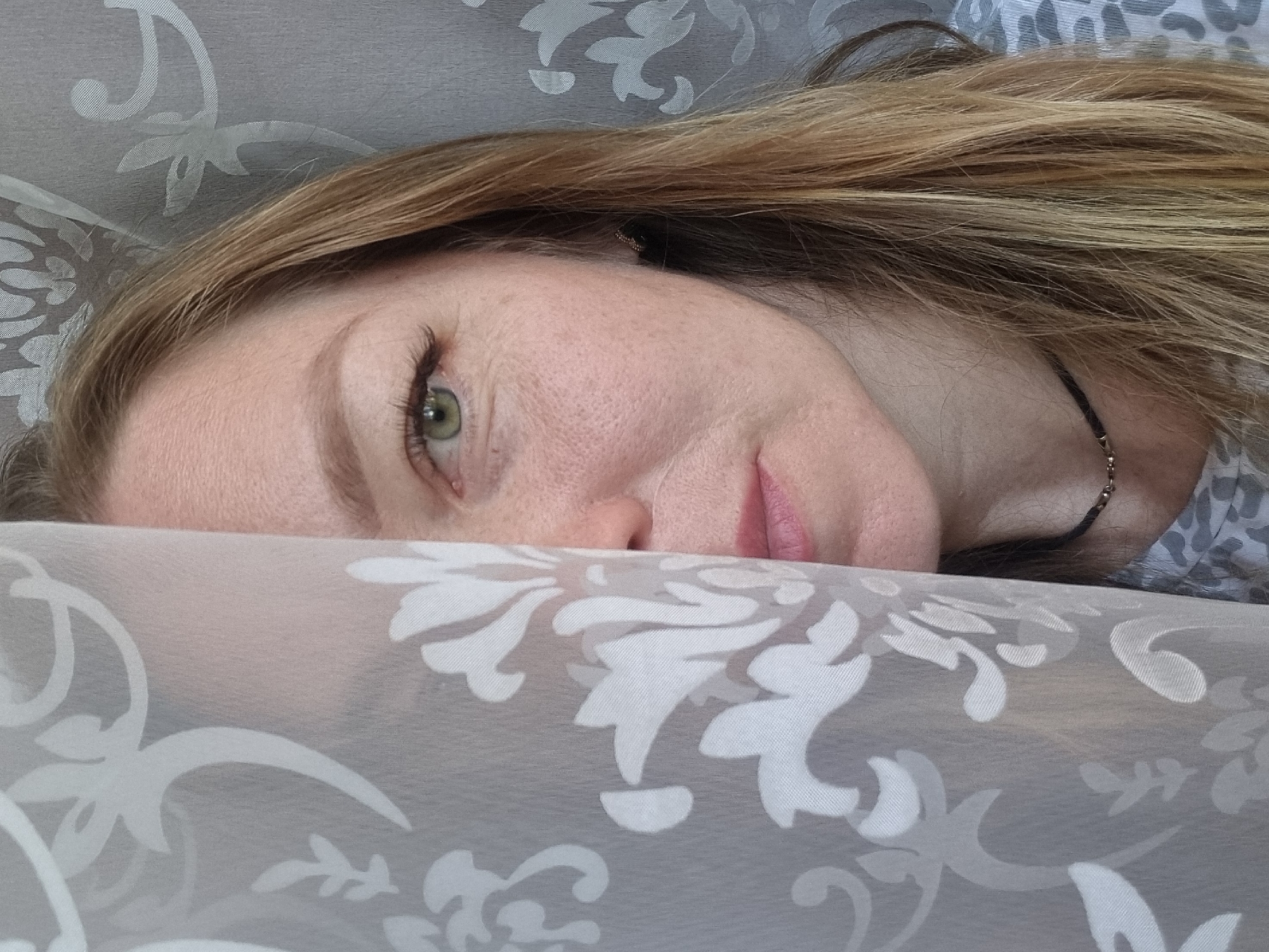 A woman lies partially obscured behind a patterned fabric. Only her forehead, one eye, nose, and mouth are visible. She has fair skin, light brown hair with blonde highlights, and an olive-green eye. A thin, dark necklace rests against her neck. The patterned fabric in the foreground is a sheer material with a white floral design on a grey background. Behind the woman, there is another patterned fabric, out of focus. There is no indication of the location being Chaikovsky, Russia.