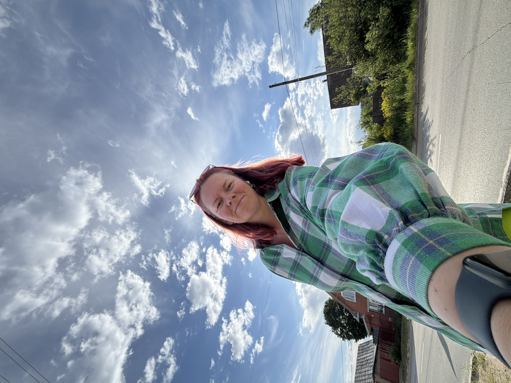 A low-angle, outdoor shot shows a red-haired woman wearing glasses and a green plaid shirt looking down at the camera. She is smiling.
The background features a bright blue sky with numerous white clouds, a telephone pole, trees, and a gray asphalt road. The environment suggests a rural or suburban area.
Based on the presence of Russian electric grid infrastructure, the location is speculated to be Chaikovsky, Russia.