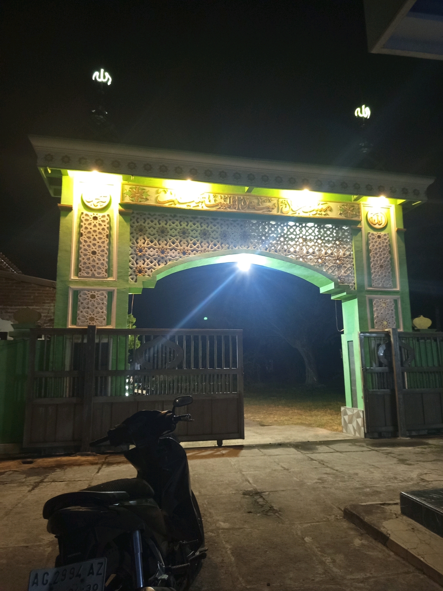 The image shows a mosque gate at night in Ngablak, Indonesia. The gate is illuminated by yellow lights and decorated with Islamic calligraphy and geometric patterns. A dark-colored motorcycle is parked in front of the gate. The ground is paved with concrete, and a metal fence is visible to the left and right of the gate. The sky is dark, suggesting it is late at night.