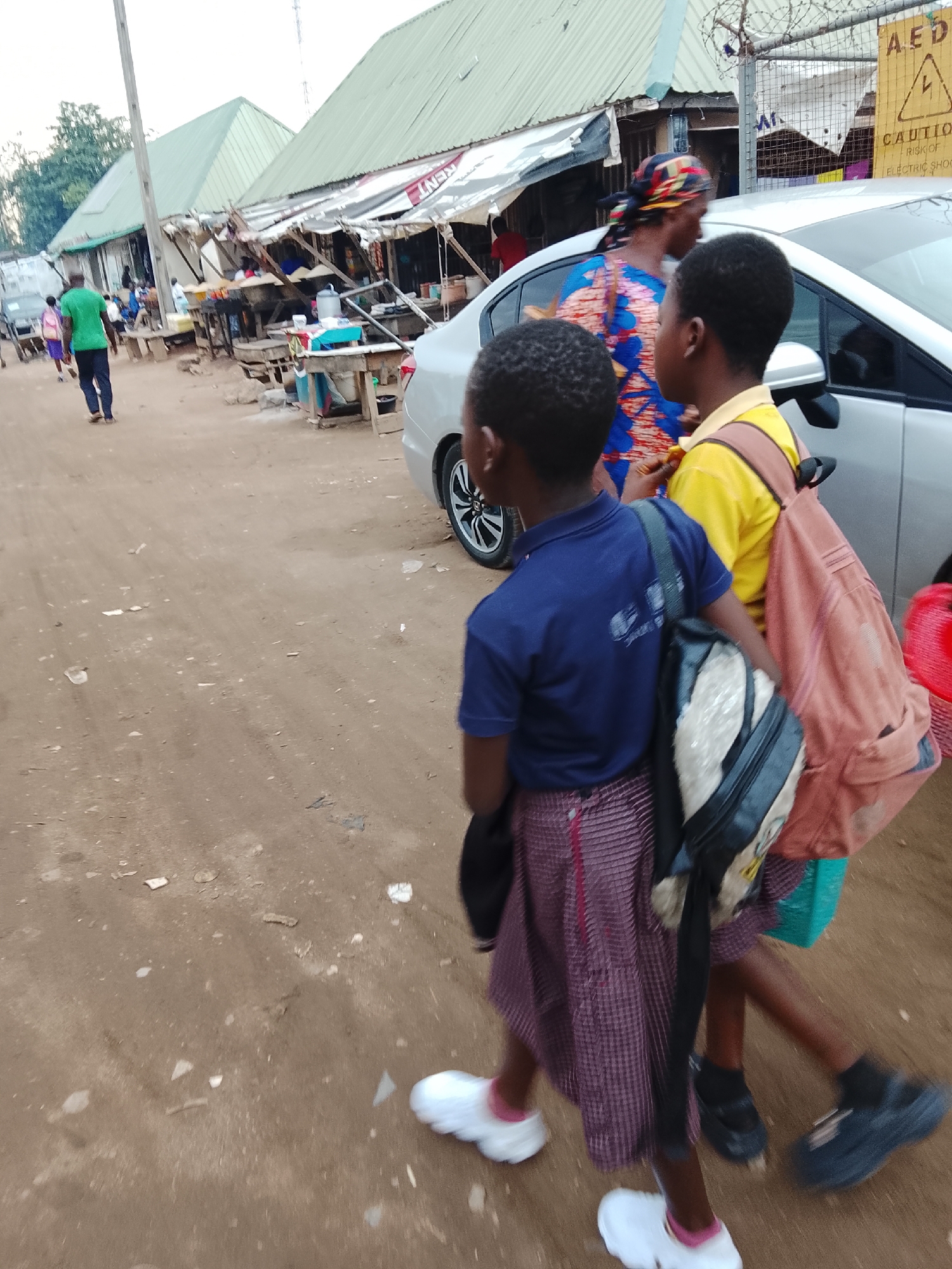 The image captures a street scene in Abuja, Nigeria, with two young people in the foreground, walking away from the camera. The person on the left wears a blue polo shirt and a checkered skirt, carrying a backpack. The person on the right wears a yellow polo shirt and a backpack, and is partially obscured by the person in front. Both are wearing dark socks and casual shoes.
Behind them, a silver car is parked. The background features a row of market stalls with green corrugated roofs. Some stalls appear to be selling goods, with various items displayed on tables and shelves. A woman in a colorful headwrap and patterned dress is visible near the stalls, walking in the same direction as the children. Another person in a green shirt is seen further down the street, also walking away.
The ground is unpaved and dusty, typical of a market area. The lighting suggests it is either early morning or late afternoon, with soft shadows. A yellow caution sign with black lettering is visible on a metal fence in the upper right corner. The sign displays a warning symbol and text that appears to say "CAUTION RISK OF ELECTRIC SHOCK." The overall atmosphere is that of a bustling local market with everyday activities taking place.