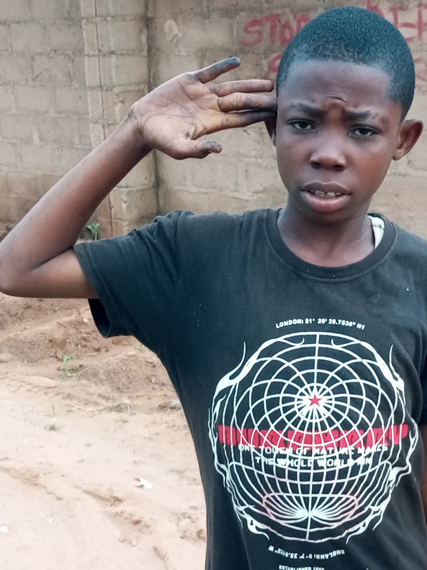 A young boy stands in what appears to be a dirt yard with a brick wall in the background. He is wearing a black t-shirt with a graphic design of a globe and text. The boy is raising his right hand to his forehead in a gesture that resembles a salute. His expression is neutral, and he is looking directly at the camera. The environment appears to be outdoors, with loose dirt and some sparse vegetation visible. The brick wall behind him has some red graffiti.