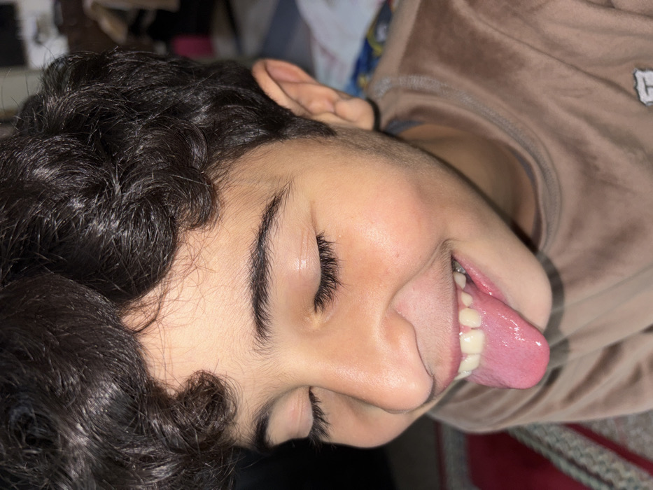A person lies on their side with their eyes closed and tongue sticking out. Dark, curly hair surrounds the face, contrasting with the lighter skin tone. The person wears a light brown, velvety top with a logo visible on the collar. The background is softly blurred, showing indistinct shapes and colors.