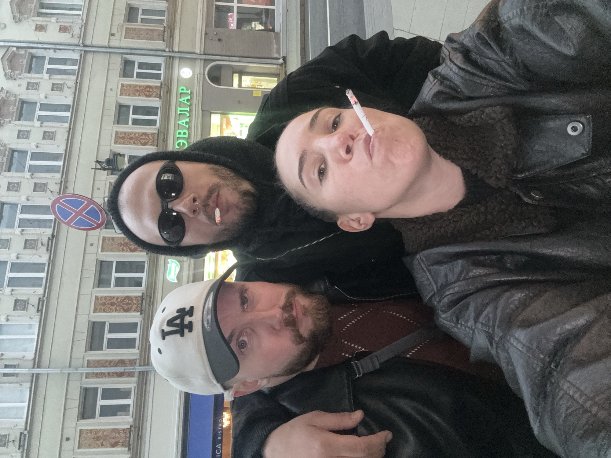 Three individuals pose for a selfie on a Moscow street. The person on the top left wears a black beanie, sunglasses, and a black jacket. They have a cigarette in their mouth. The person on the top right is also holding a cigarette in their mouth and wears a black leather jacket with a fur collar. The person at the bottom wears a baseball cap and a leather jacket. Behind them, a building with multiple windows and a green sign with "ЭВАЛАР" is visible. A street sign with a red cross is also visible near the upper left person.