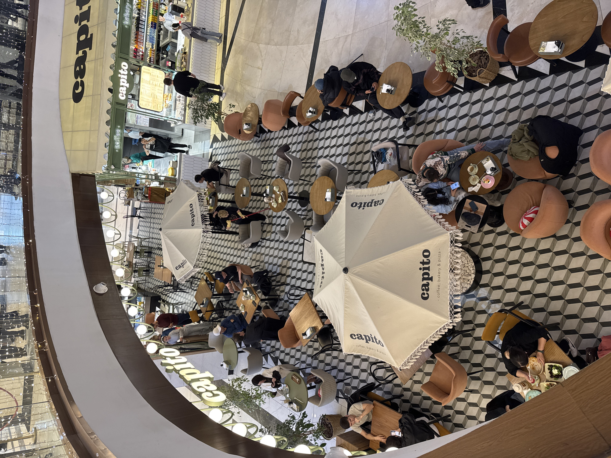 This overhead shot captures a lively cafe named "capito," situated within what appears to be a multi-story indoor shopping mall or commercial complex. The view is from an upper level, looking down onto a sprawling seating area with a distinctive black, white, and grey geometric tiled floor, creating a 3D cube illusion.
Numerous individuals are scattered throughout the cafe. Most are seated at a variety of tables and chairs, including comfortable upholstered armchairs in shades of brown and grey, and simpler wooden and metal seating. People are engaged in casual activities such as eating, drinking, conversing, and using mobile phones. Plates with food, cups, and personal belongings are visible on many tables. Several staff members are seen, both behind a service counter and moving through the seating areas.
Two large, cream-colored umbrellas with the "capito" logo and slogan are strategically placed among the tables, creating a defined ambiance. Green potted plants add a touch of nature to the indoor setting. The "capito" brand is prominently displayed in large, illuminated letters on a facade, and also on the umbrellas and above the counter area.
Visible text includes:
- "capito" (multiple instances, including a large illuminated sign and on umbrellas)
- "coffee, bakery & pizza" (on umbrellas and above the counter)
- "coffee bakery & pizza" (on umbrellas)
- "-bakery-" (above the counter).
The scene suggests a relaxed, social atmosphere, likely during business hours, well-lit by interior lighting.