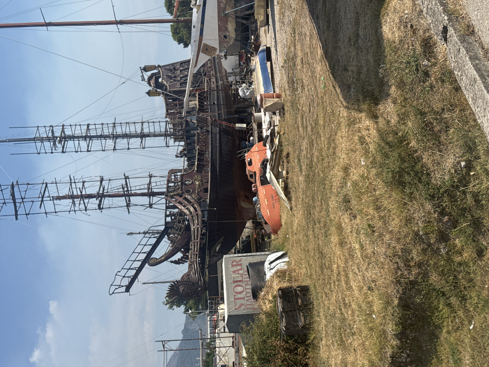 The image shows a dry, grassy outdoor area with several boats and marine structures under construction or repair. In the background, a large, old-fashioned sailing ship with multiple masts and scaffolding is prominent. To the right of this ship, a white boat with a blue stripe is propped up on stands. In the foreground, an orange life raft is visible, along with various discarded materials and wooden crates. A grey container with Russian text, "СТОЛАР ПЛОТНИК," is also present. The environment suggests a shipyard or a maritime repair facility in Bar, Montenegro, with dry vegetation and a clear blue sky overhead.