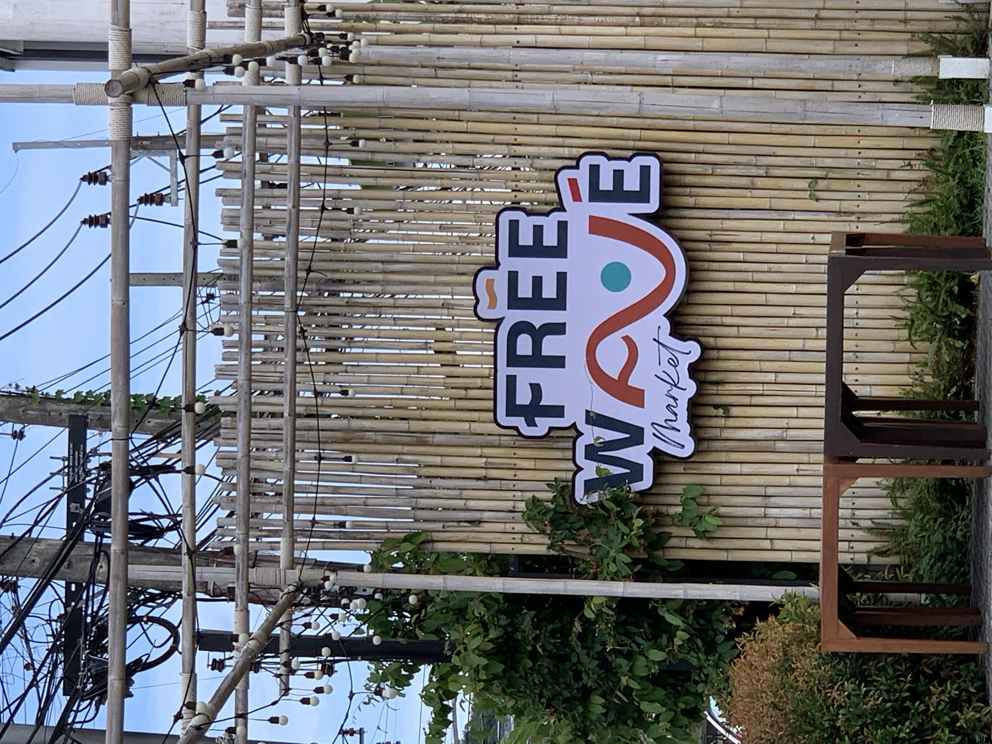 This image depicts an outdoor market entrance in Baan Bo Phut, Thailand, featuring a prominent sign set against a bamboo structure. The central element is a large, oval-shaped sign with a white background and a dark gray outline, displaying the text "FREE WAVE Market". "FREE" is rendered in bold, dark gray sans-serif font, with a small orange accent mark above the first 'E'. Below it, "WAVE" is in a similar dark gray font, but the 'A' is stylized as an orange wavy line incorporating a teal dot, and the 'E' has an orange accent mark. "Market" is written in a smaller, cursive, white script with a dark gray outline, placed below "WAVE".
The sign is mounted on a vertical bamboo wall or fence, constructed from numerous light to medium yellow-brown bamboo poles. Above this wall, an overhead canopy framework of horizontal bamboo poles supports multiple strands of string lights, each with spherical white bulbs, which also drape downwards. Several vertical bamboo poles, lighter in color, act as structural supports and are wrapped with white rope at various intervals.
In the background on the left, multiple utility poles are visible against a light blue sky, heavily laden with a dense, chaotic network of black electrical and communication wires. Brown ceramic insulators are discernible on these poles. Green foliage and vines are intertwined with the utility infrastructure on the left side.
In the foreground, two rectangular wooden tables or low benches are situated on a dark paved surface. The table on the left exhibits a lighter brown hue, while the one on the right is a darker brown. Surrounding the base of the bamboo wall and extending into the foreground are various green bushes and plants, including a prominent bush with reddish-brown foliage on the far left. The entire scene is illuminated by natural daylight.