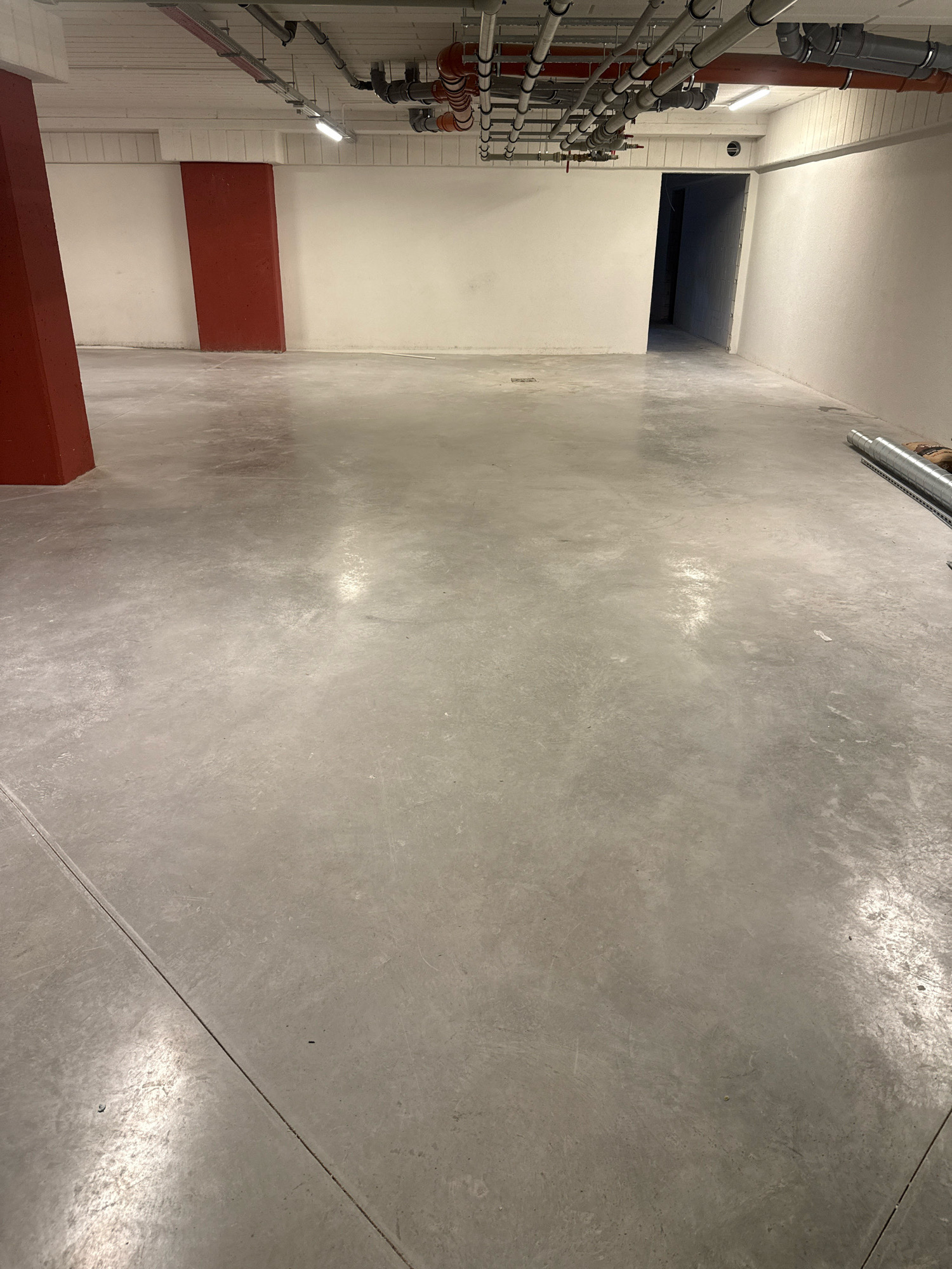 The image depicts the interior of an unfinished or industrial space. The floor is a smooth, grey concrete surface with visible expansion joints. On the left side of the image, a tall, red, rectangular pillar is partially visible. Towards the back of the space, on the left, another red pillar is visible. The walls are painted white. The ceiling features exposed piping and electrical conduits, with fluorescent lighting fixtures. In the background, an open doorway leads into a darker area. On the right side of the floor, a length of flexible silver ducting is laid out. The scene appears to be located in Nowa Sól, Poland, as per the provided context, likely within a building under construction or a utility area. There are no people or animals visible in the image.