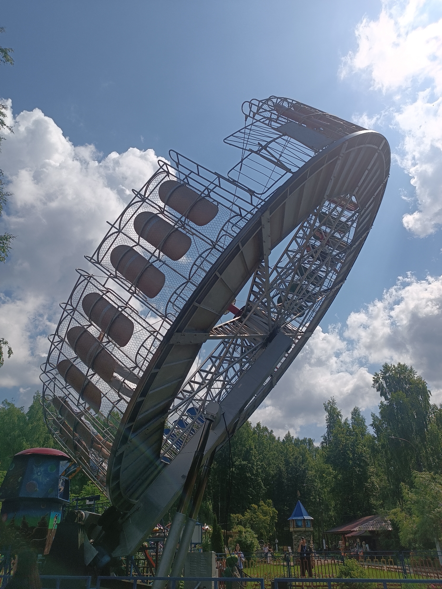 This is a medium-angle, eye-level outdoor photograph taken on a sunny day with scattered clouds, showing a large amusement park ride in the city of Chaikovsky, Russia. The ride, a large metal disc-shaped structure with rows of barrel-like seats, is tilted at an extreme angle, appearing to be in motion. The structure is predominantly gray metal with some brown seats and a red accent visible within the ride's mechanism. The foreground shows a portion of a blue fence and some greenery. In the background, a dense line of green trees forms the horizon. At the base of the amusement ride, to the left, is a colorful, cartoonish structure resembling a spaceship or rocket, with blue, red, and black elements decorated with cosmic imagery. Further in the background, several people are visible, as well as a small building with a dark roof and a blue-capped tower structure. The sky is a vibrant blue with fluffy white clouds.