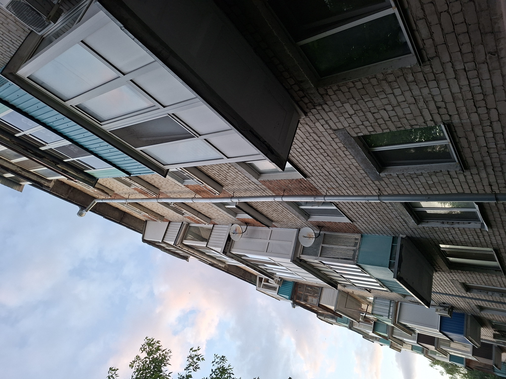 The image shows a low-angle shot of a brick apartment building in Kazan, Russia. The building has multiple balconies, many of which are enclosed with glass and have turquoise accents. A downspout runs vertically along the side of the building. The sky above is partly cloudy with hints of pink and blue. A few trees are visible on the left side of the frame. The scene appears to be an exterior shot of residential housing.