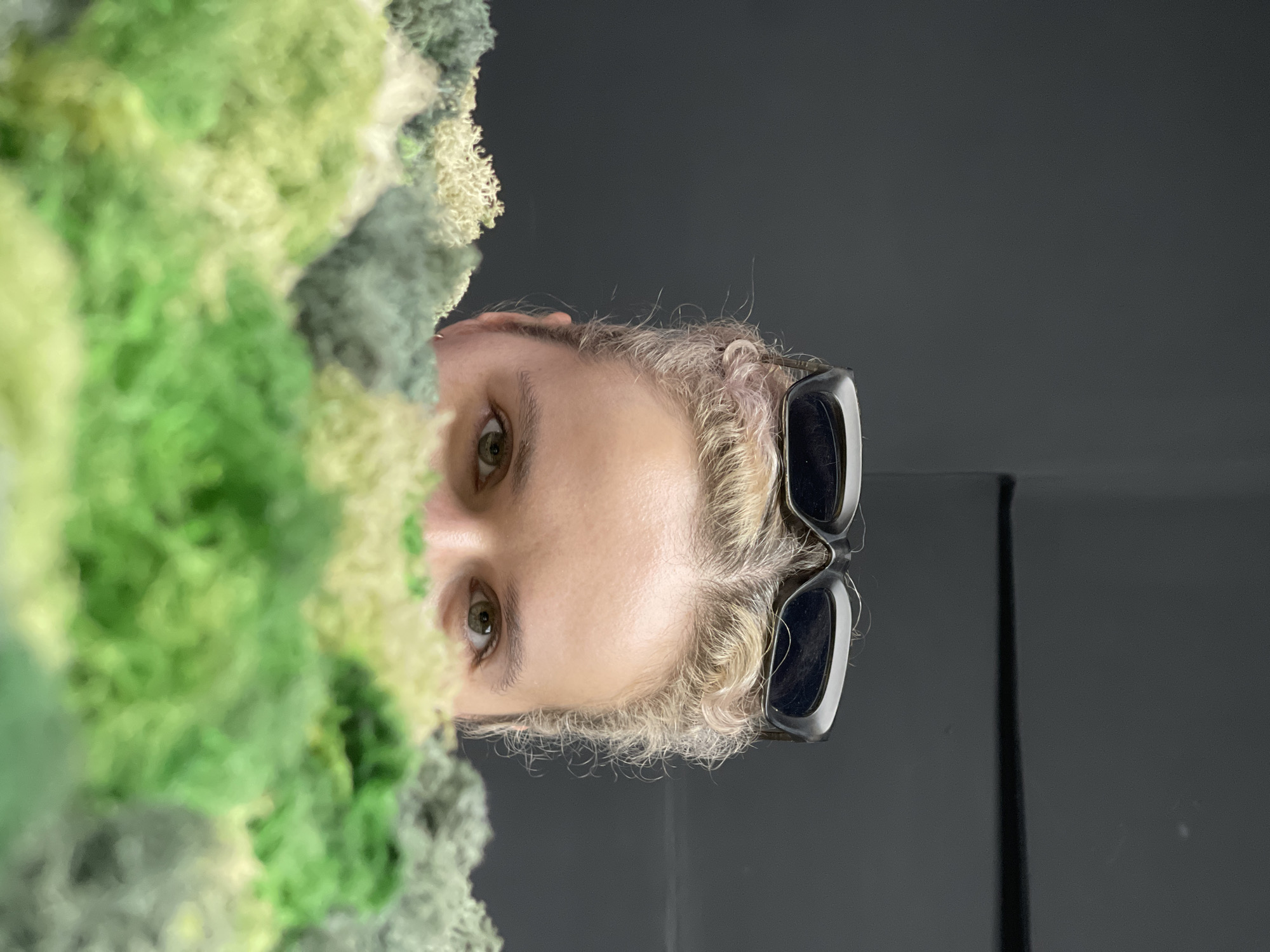 A close-up, eye-level shot, taken in Thủ Đức, Vietnam, features a person's upper face partially obscured by a dense, textured mass of green and yellow moss on the left. The individual, with light blonde or bleached hair and dark sunglasses pushed up onto their head, gazes directly forward with a neutral to slightly inquisitive expression. Their eyes are light-colored, possibly hazel or green. The moss, appearing soft and organic with varying shades of vibrant green and lighter yellowish-green, fills the foreground on the left, creating a natural, somewhat blurry barrier. The background on the right and behind the person is a plain, dark grey or black surface, suggesting an indoor setting or controlled environment. There is no visible text in the image.