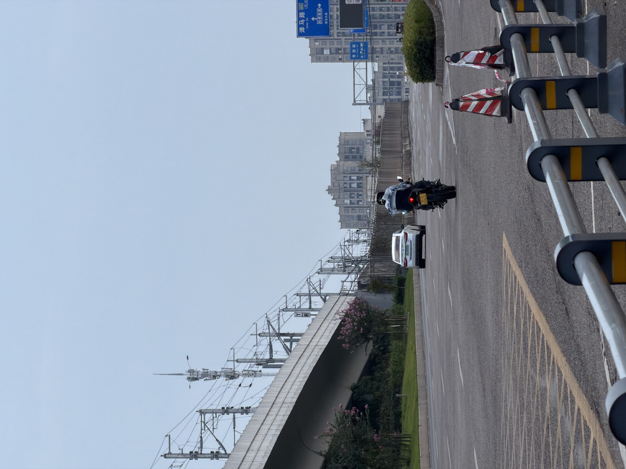 A motorcycle rider, wearing a blue jacket and white helmet, is seen from behind on a road in Jiangjin, China. The rider is traveling away from the camera. To the left of the road, a white car is also traveling in the same direction. Elevated train tracks with overhead power lines dominate the left side of the frame, running over a concrete overpass. The background consists of buildings and a clear, overcast sky. Blue road signs are visible on the right side of the road, indicating directions and street names. Several traffic cones and a metal barrier are in the foreground.