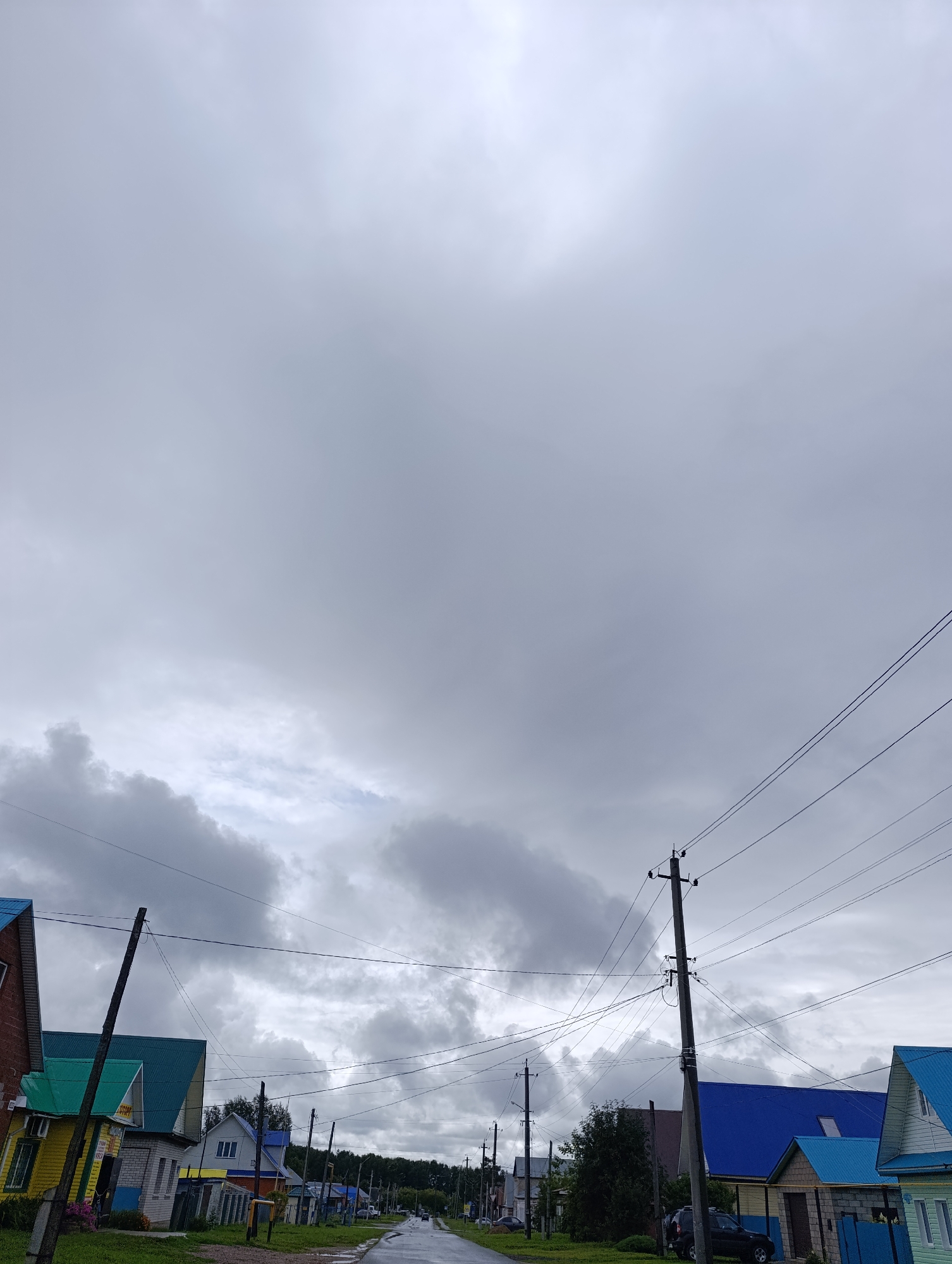 The image depicts a residential street in an unknown location, under a cloudy sky. The street is paved and wet, suggesting recent rainfall. Several houses with colorful roofs, predominantly blue and green, are visible lining the street. Power lines and utility poles crisscross the scene, indicating infrastructure typical of a populated area. Some cars are parked along the street or in driveways. The overall impression is one of a quiet, overcast day in a small town or village.