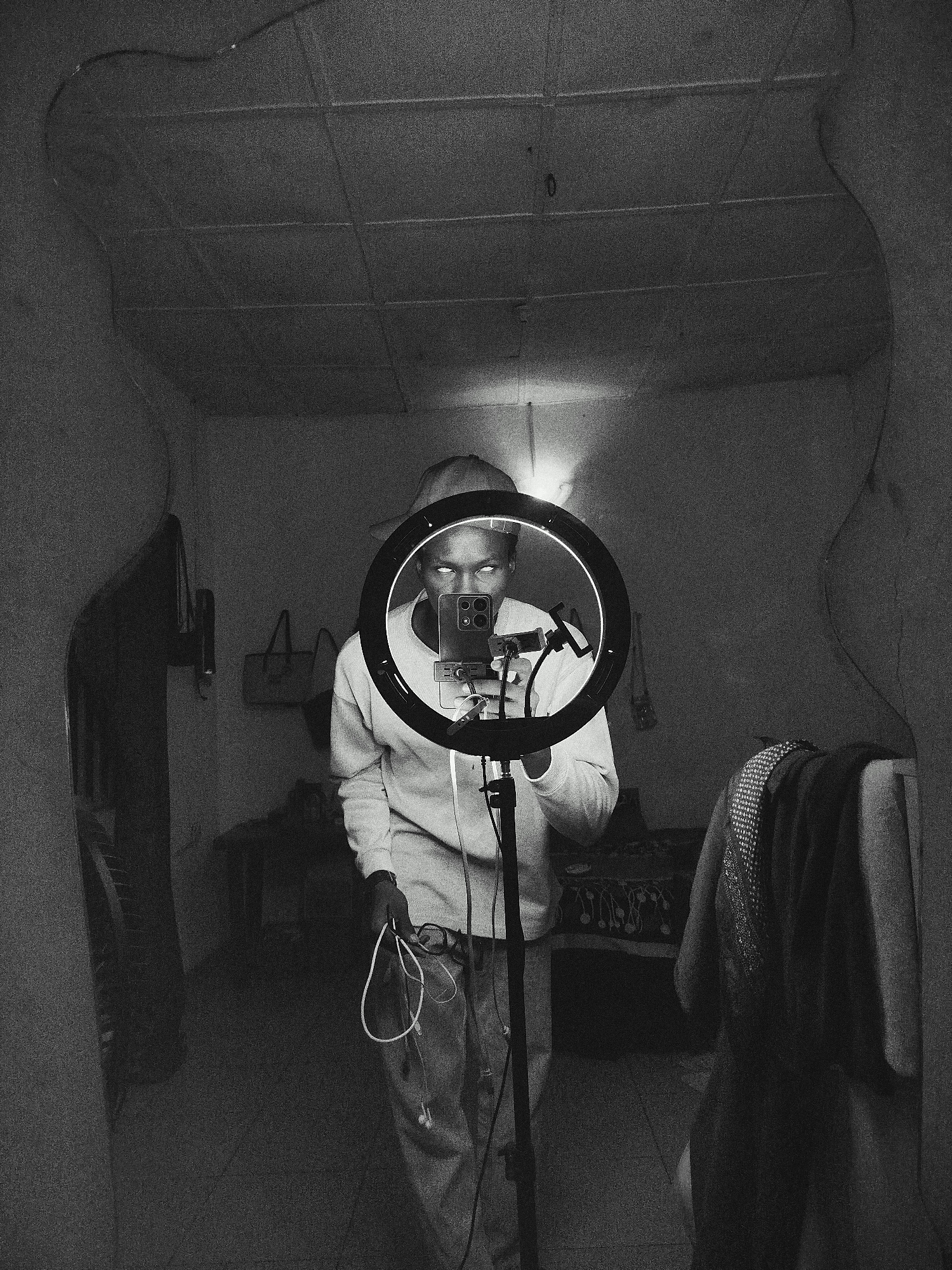 A black and white photograph shows a person reflected in a mirror holding a cell phone mounted on a ring light. The individual is standing in what appears to be an indoor setting with clothing and household items visible in the background. The mirror's irregular shape frames the scene, reflecting a ceiling with patterned tiles. The person holds wires or cables in one hand and is wearing a long-sleeved shirt and pants.