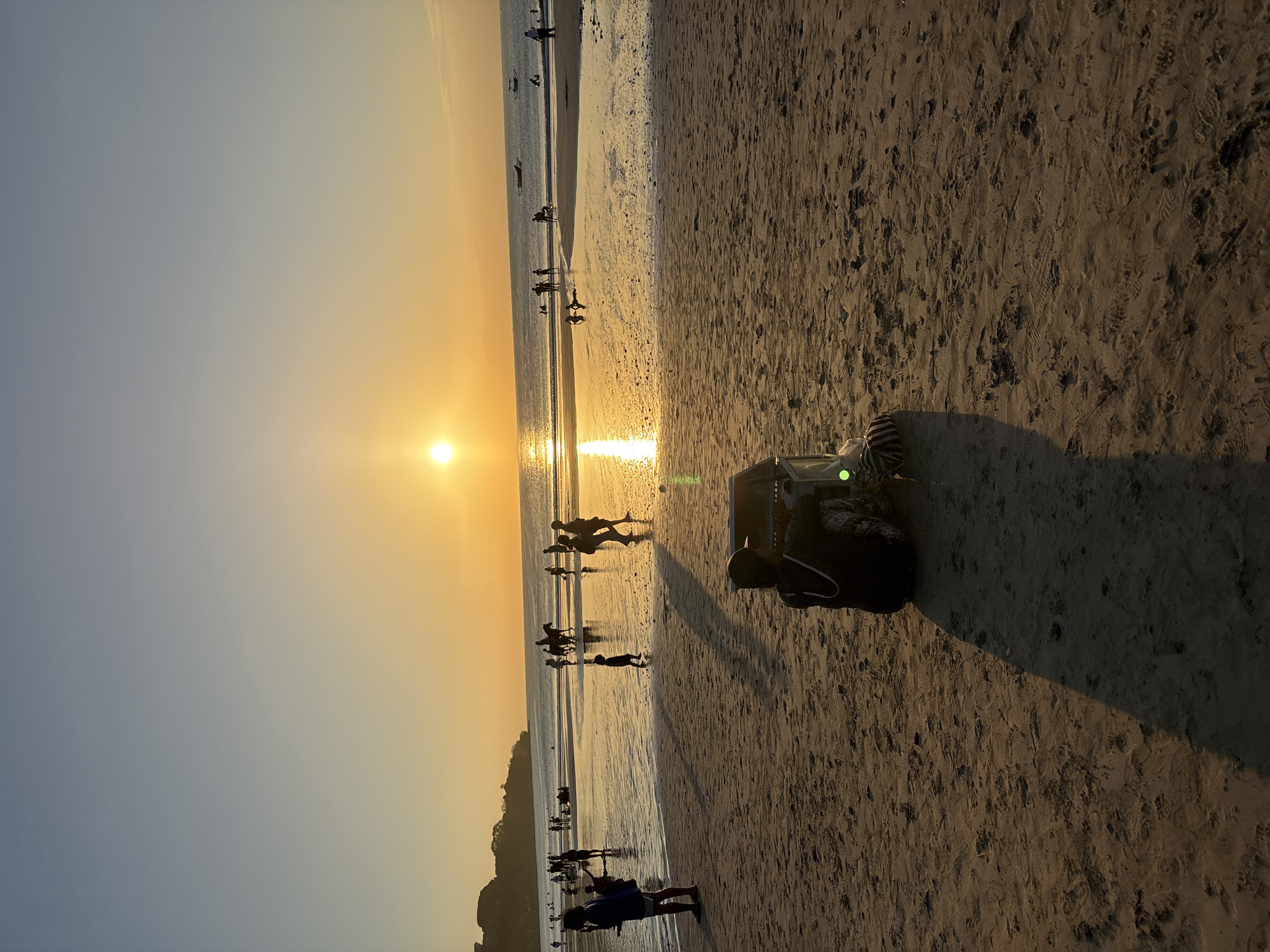 A wide shot captures a beach scene at sunset in Kuta Selatan, Indonesia. The sun, a bright orb low in the sky, casts a warm golden glow across the water and sand. Numerous people are scattered across the beach and in the shallow water, some walking, others sitting, and a few riding horses. The foreground features a person sitting on the sand near a small cart filled with items. The sand is marked with footprints. The ocean extends to the horizon, with a few boats visible in the distance. The sky transitions from a pale blue at the top to a warm orange near the horizon.