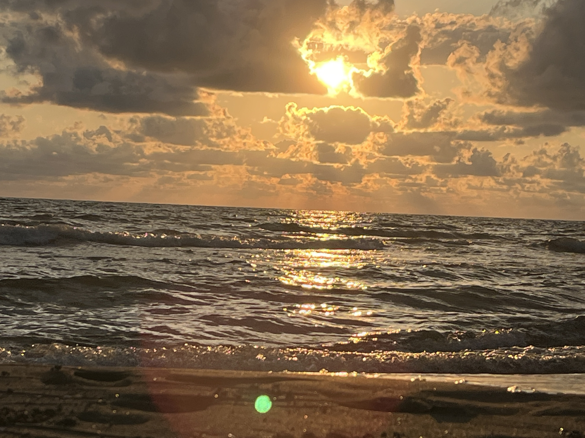 The image depicts a sunset over the Black Sea. The sun is partially obscured by dramatic, golden-orange clouds, casting a shimmering path of light across the rippling water. The foreground shows the wet sand of a beach, with a faint green bokeh effect in the lower part of the frame. There are no people or man-made structures visible, suggesting a natural and tranquil scene. The location is specified as Sufsi Community, Georgia.