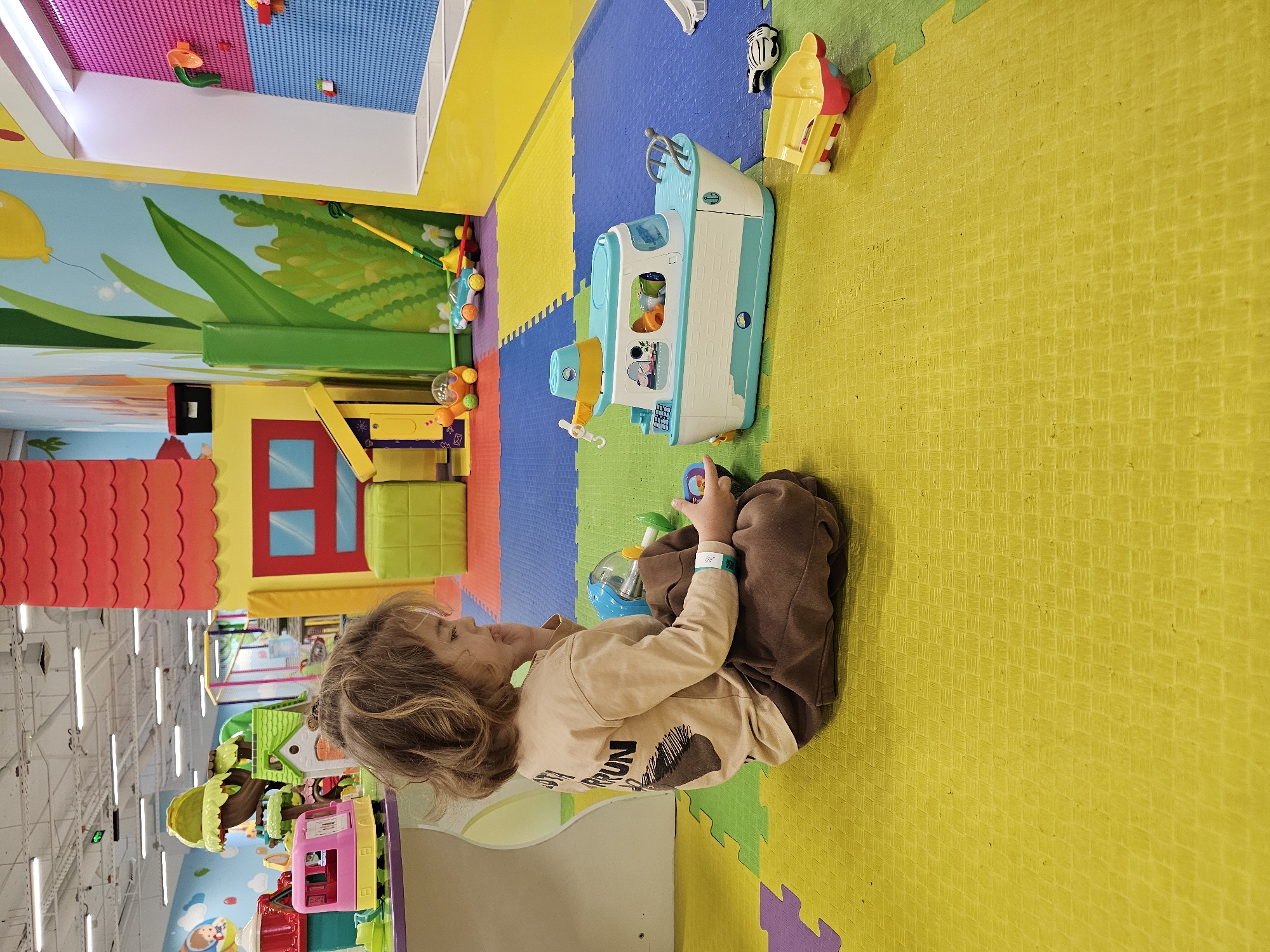 A child is sitting on a yellow and green padded play mat in a brightly colored indoor play area, possibly in Khimki, Russia, based on visual elements. The child, with curly hair, is wearing a beige shirt and brown pants. The child is playing with a toy boat. Other toys are scattered nearby, including a zebra toy and a toy ice cream cone. The play area is decorated with colorful murals and various play structures, including a red-roofed playhouse, suggesting a children's entertainment center. The lighting appears to be artificial and bright.