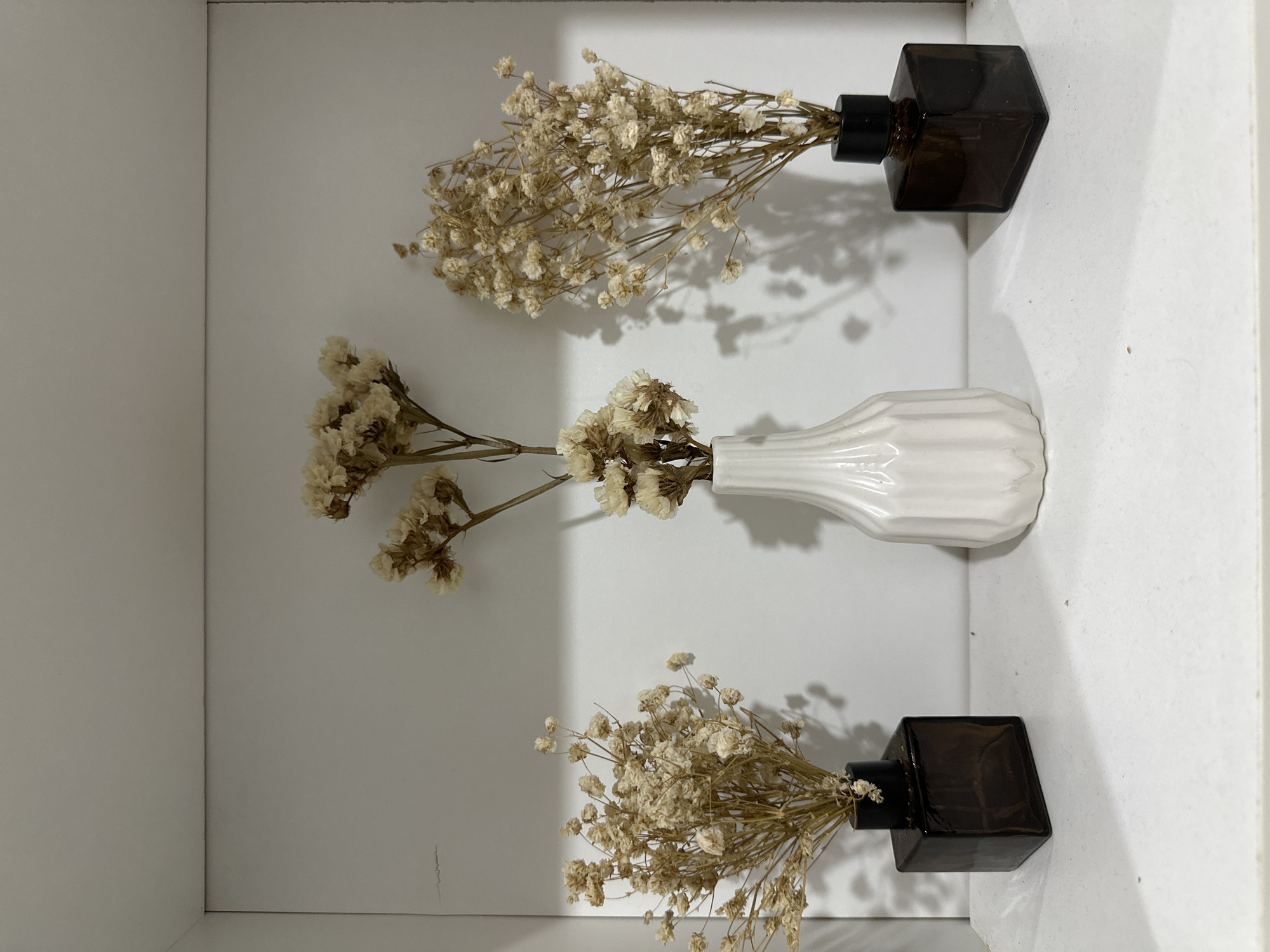 The image shows three vases of dried baby's breath flowers arranged on a white shelf, likely inside a building. The top and bottom vases are identical dark brown, square bottles with black stoppers, while the middle vase is white and ribbed. The flowers are dried and a light tan color. The shelf appears to be part of a built-in unit or cabinet, with white walls on the left, top, and right sides. There is no evidence in the image to suggest it is located in Davao City, Philippines. There are no people visible.