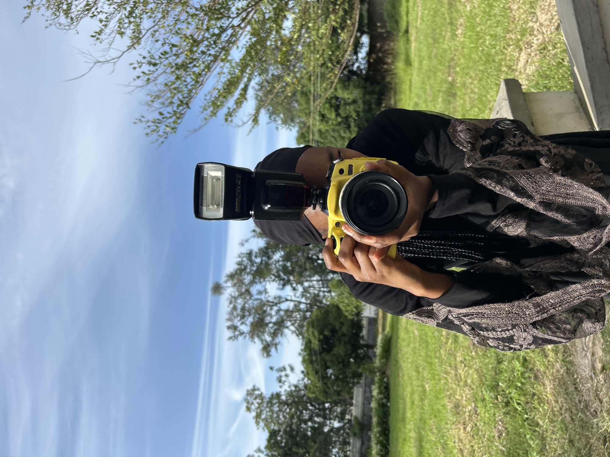 A person wearing a black hijab and a black garment with ornate brown detailing on the sleeves is holding a yellow camera with a flash attachment. The person is looking through the camera, which is pointed directly at the viewer. The background consists of a grassy field, trees, and a blue sky with wispy clouds. The scene appears to be outdoors in a semi-rural or park-like setting. The composition of the image suggests the person is taking a photograph or posing for one. The lighting indicates it is daytime. The location is stated to be Vom, Nigeria.