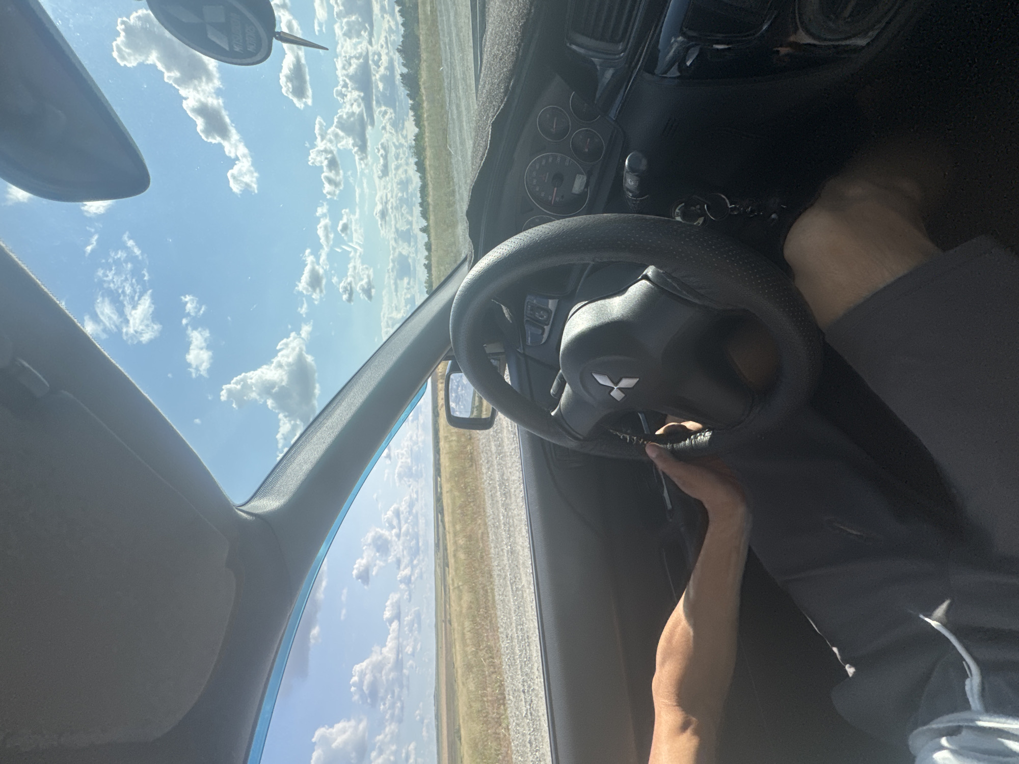 The image shows the interior of a car driving on a dirt road through a rural landscape. A person's arms are visible, one resting on the steering wheel, which has a Mitsubishi logo. The dashboard and part of the windshield are also visible, with a clear sky and clouds above. The landscape outside consists of dry grass and some distant trees. The scene is bathed in sunlight. There is no indication of the location being Васильевка, Russia.