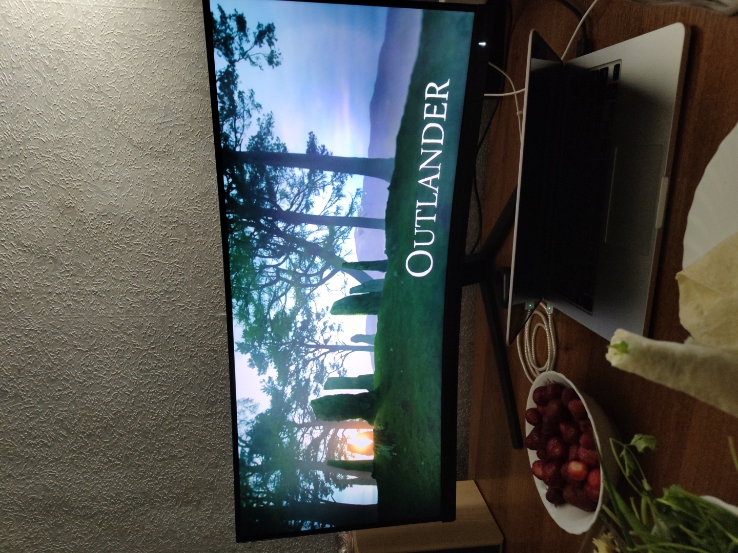 A widescreen computer monitor displays the title "OUTLANDER" in large white text against a background of silhouetted trees and standing stones at sunrise. In front of the monitor, on a wooden desk, rests a partially opened laptop. To the left of the laptop is a white bowl filled with fresh strawberries. To the left and foreground, there is a white plate with a partially wrapped tortilla and some fresh greens, possibly cilantro or parsley, in a white bowl. A charging cable is connected to the laptop and is coiled on the desk. The scene appears to be indoors, with a textured wall visible in the upper portion of the image.