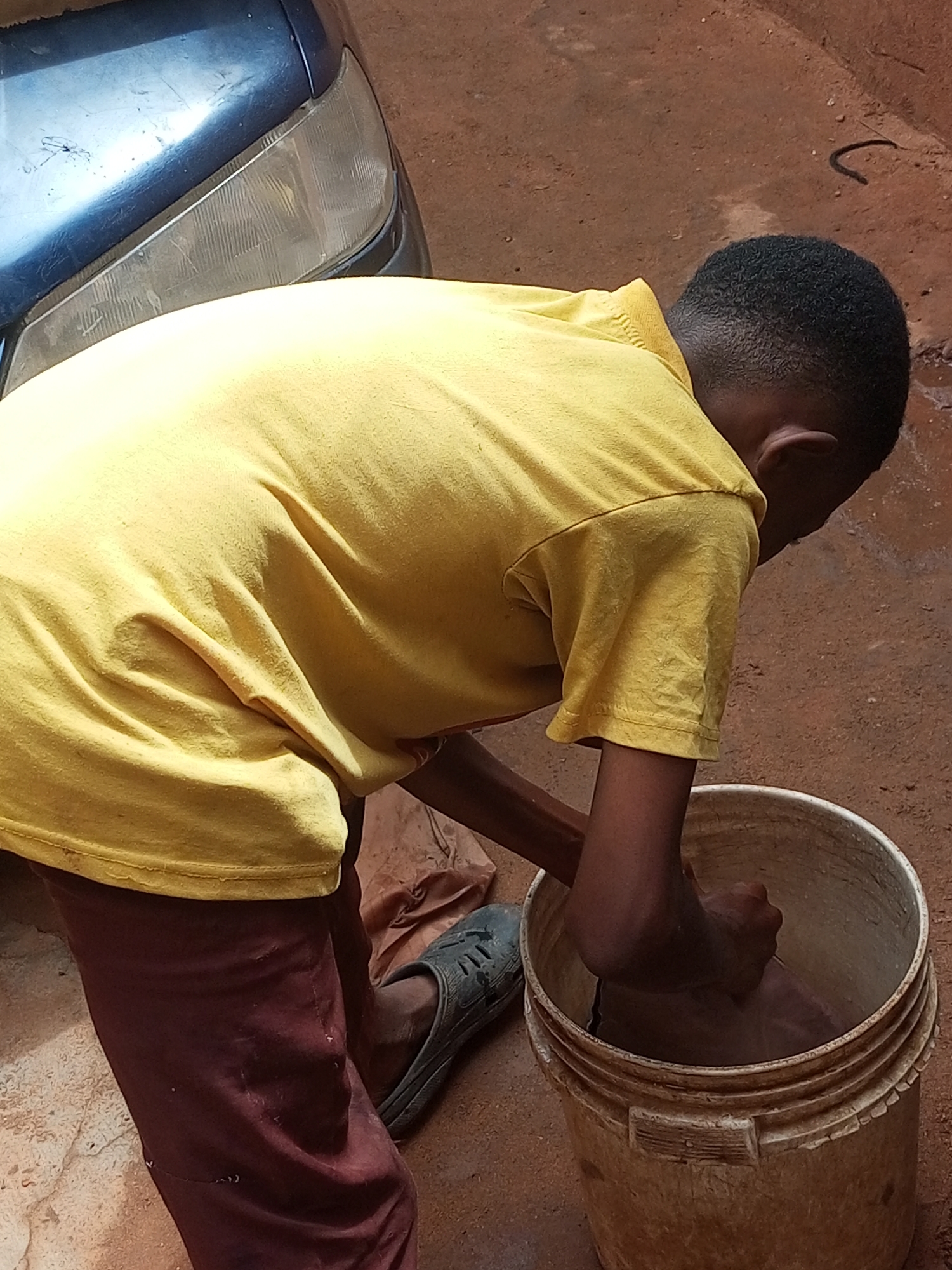 A young Black male wearing a yellow t-shirt and maroon pants is kneeling next to a white plastic bucket filled with water. His right hand is submerged in the bucket, and he appears to be washing something. His left leg is bent and his foot is also inside the bucket. His dark-colored shoe is visible next to the bucket. The ground around him is a mixture of reddish-brown soil and some debris. In the background, the front of a dark-colored vehicle with a clear headlight is visible. The scene appears to be outdoors, possibly in a residential area within Gombe, Nigeria.