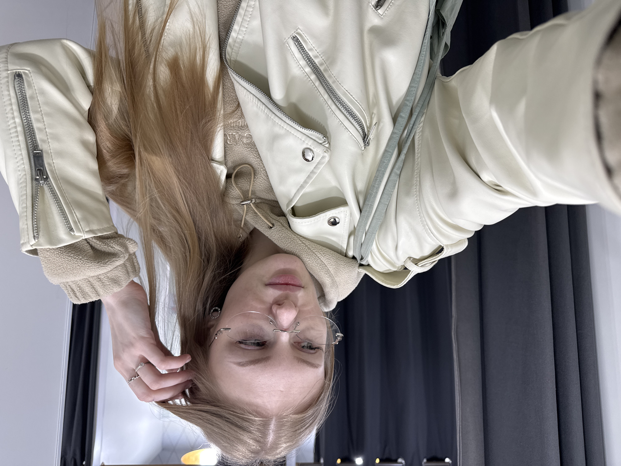 An indoor selfie captures a young individual, likely female, from the chest up. The original image is inverted, showing the person upside down. When correctly oriented, she has long, light brown to blonde hair flowing over her shoulders. She wears a cream or off-white faux leather biker jacket featuring numerous silver-toned zippers and snap buttons, layered over a beige hooded sweatshirt or top with a visible fleecy or sherpa texture on the cuffs and around the hood.
Her face is centrally placed, framed by her hair. She is wearing rimless glasses with a subtle silver-toned nose bridge. Her expression is relatively neutral, with her left eye appearing partially closed or squinting. Her right hand, adorned with a simple silver ring on the ring finger, is raised to touch her hair near her temple. A silver hoop earring and another small silver piercing are visible on her left ear.
The background is simple, featuring dark gray pleated curtains on the right side of the frame and a plain light-colored wall or surface on the left. The lighting suggests an indoor setting, and no specific time of day or weather is indicated. Indistinct, faint text, possibly "CAR", can be vaguely seen on the beige inner garment, but it is not clearly legible. The photo is taken in Perm, Russia.